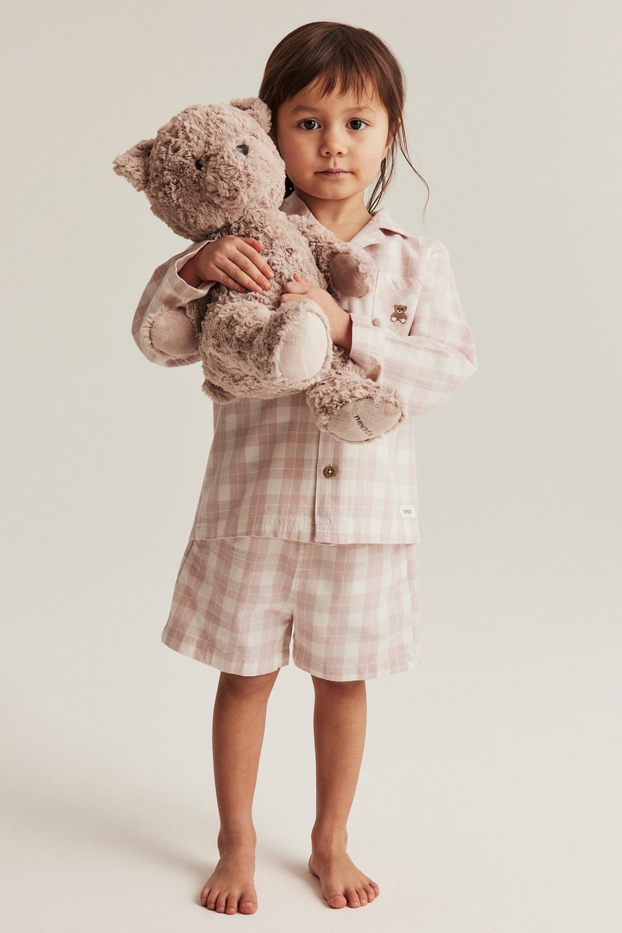 Front view of a kid wearing a pink and white checked cotton pyjama set, holding a light brown teddy bear.