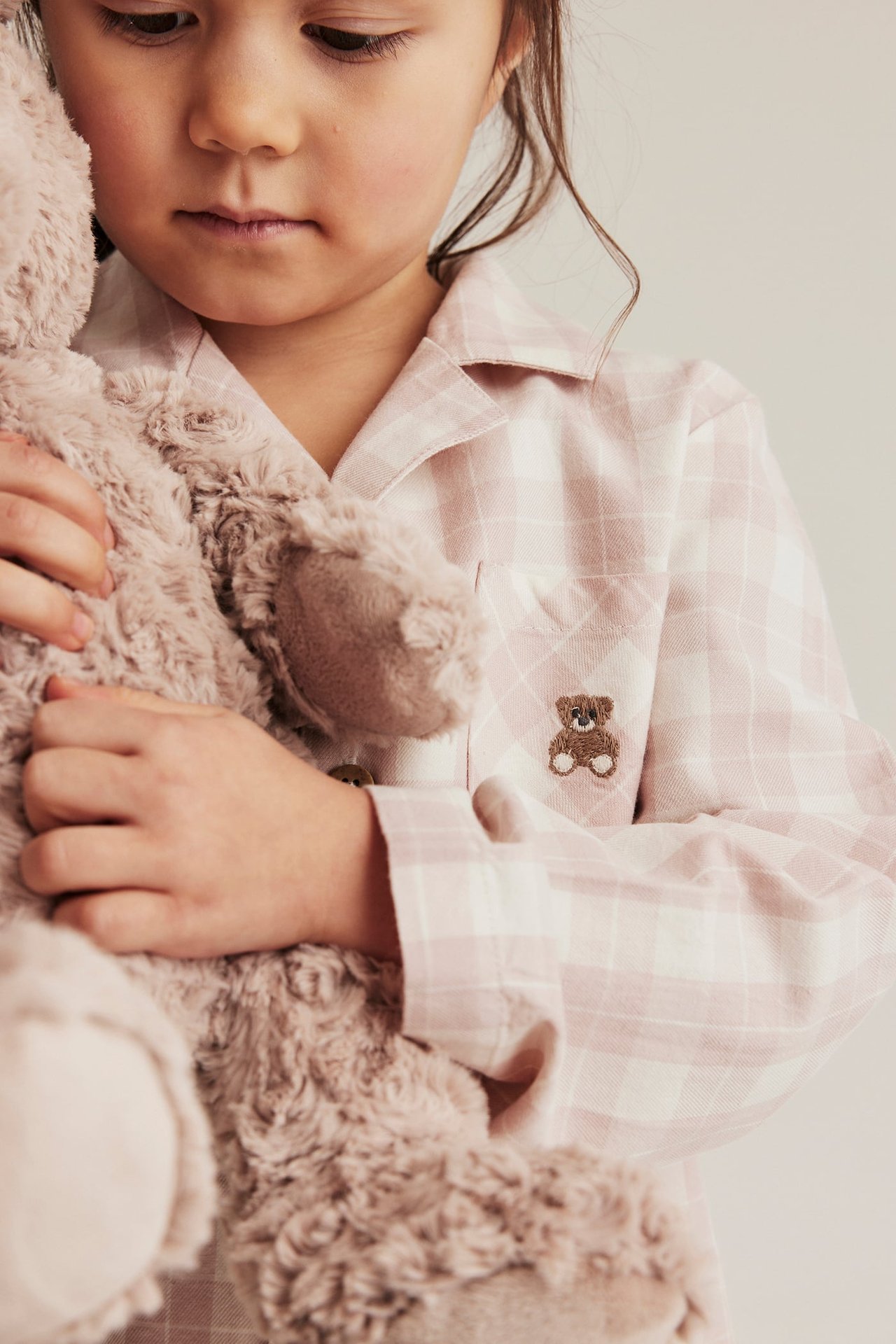 Nahaufnahme eines Kindes in rosa kariertem Baumwoll-Pyjamahemd mit Teddybär-Stickerei, hält einen flauschigen Teddybär.