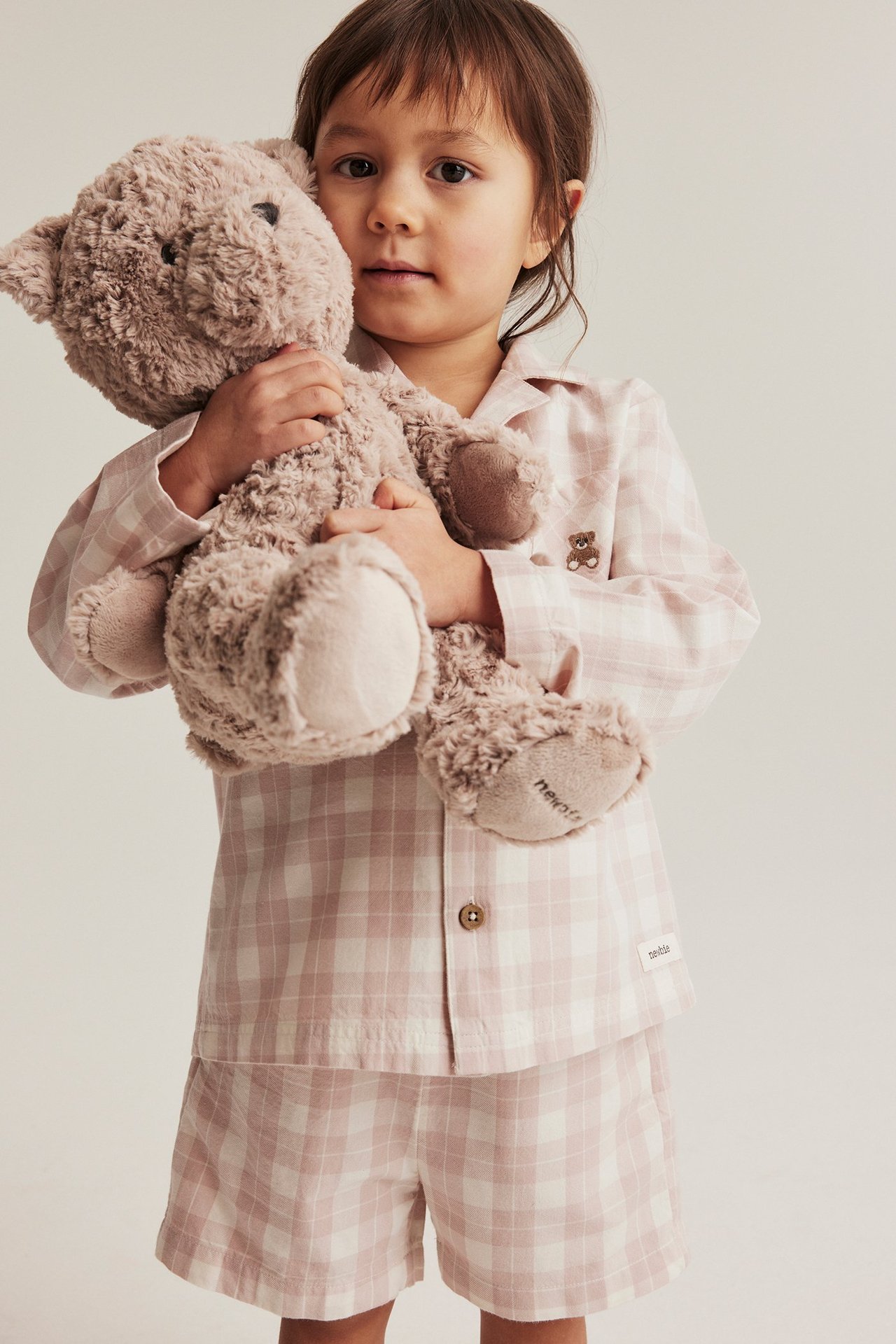 Front view of a kid wearing pink checkered cotton pyjamas, holding a brown teddy bear.