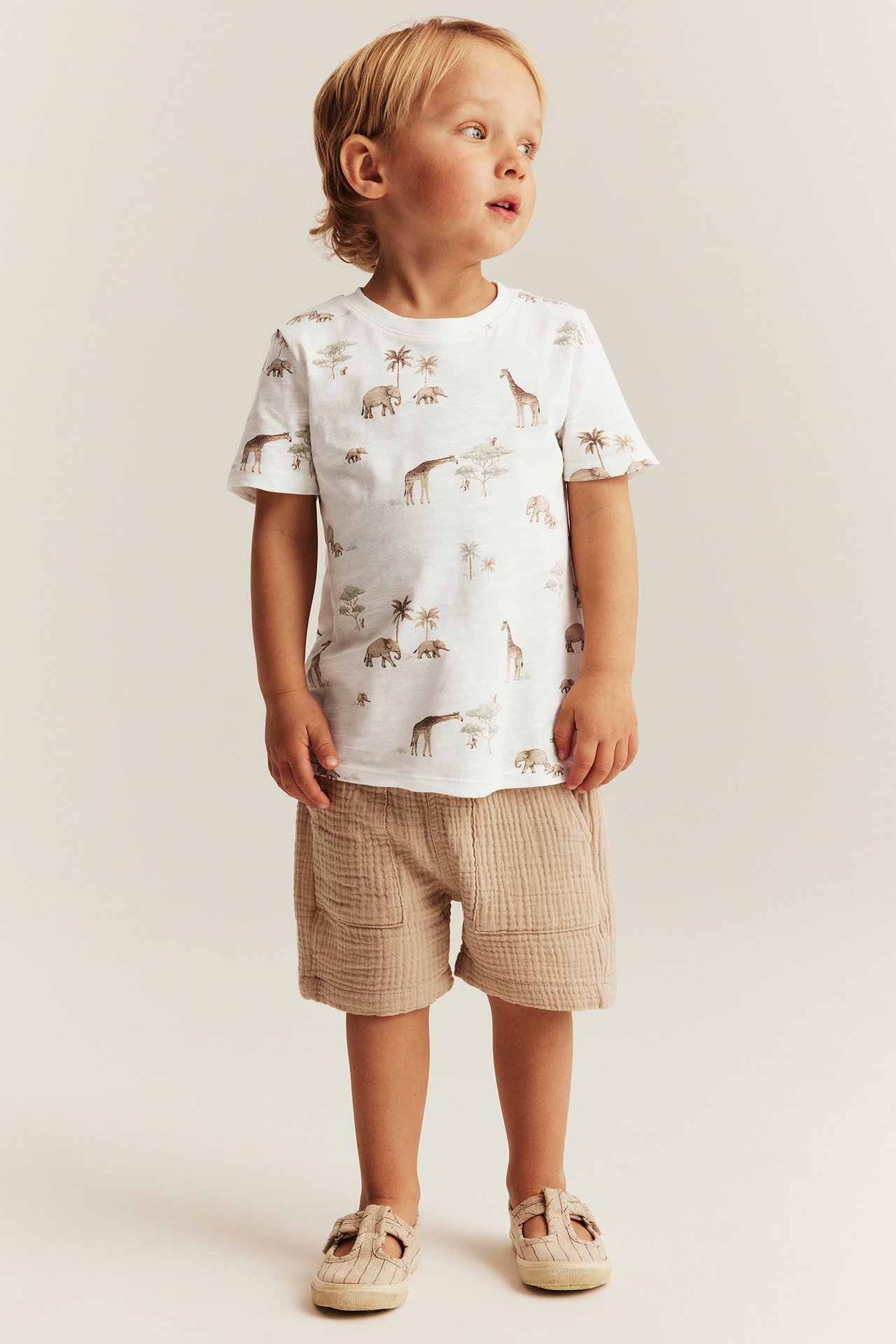 Front view of a kid wearing an off-white animal-patterned cotton t-shirt, beige textured shorts, and beige sandals.