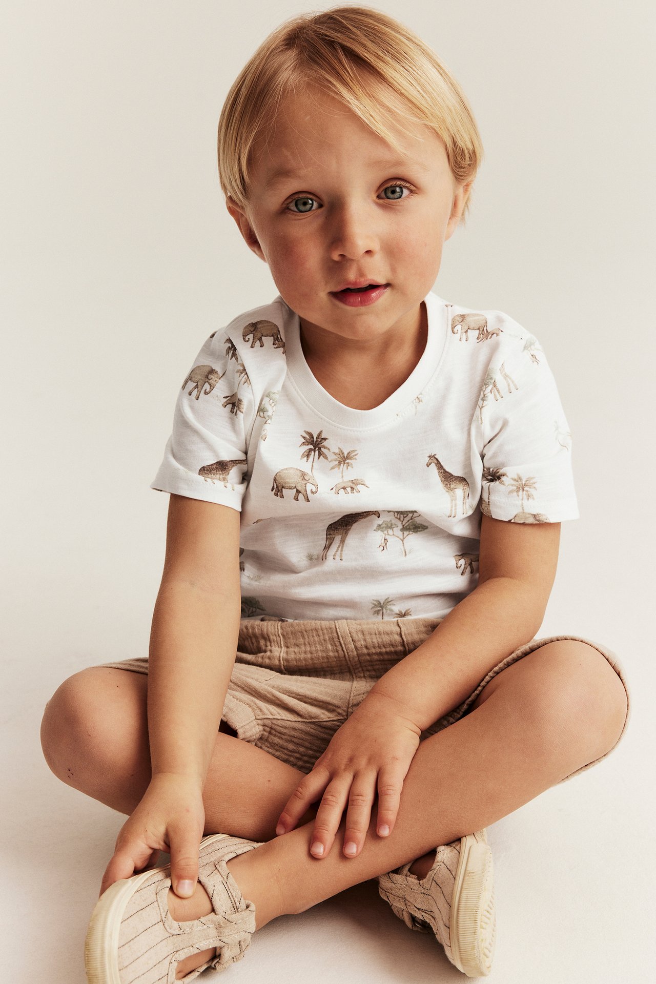Front view of a kid wearing an off-white animal print short-sleeved t-shirt, beige shorts, and striped canvas shoes.