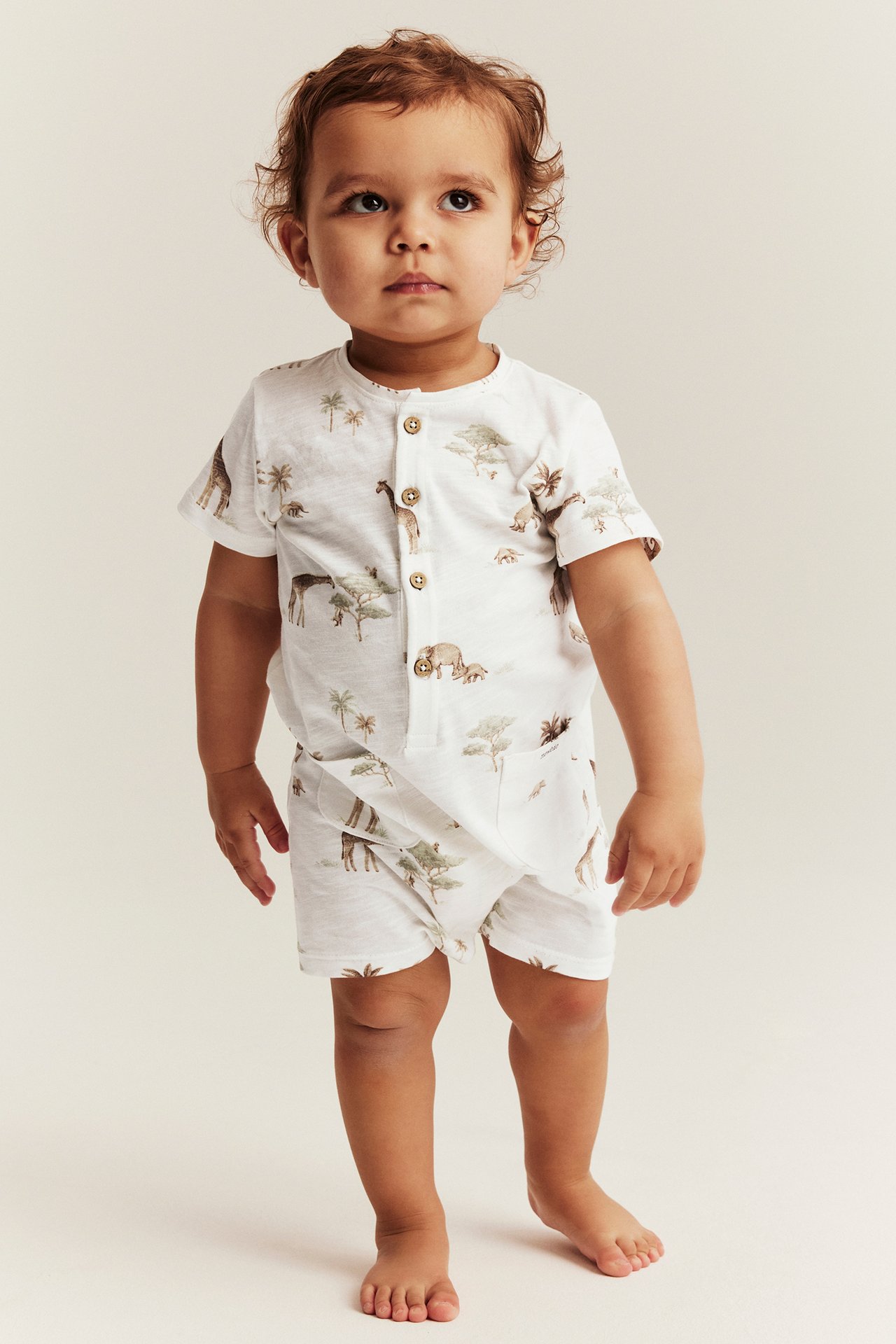 Front view of a baby wearing an off-white cotton animal-patterned romper with front buttons and patch pockets.