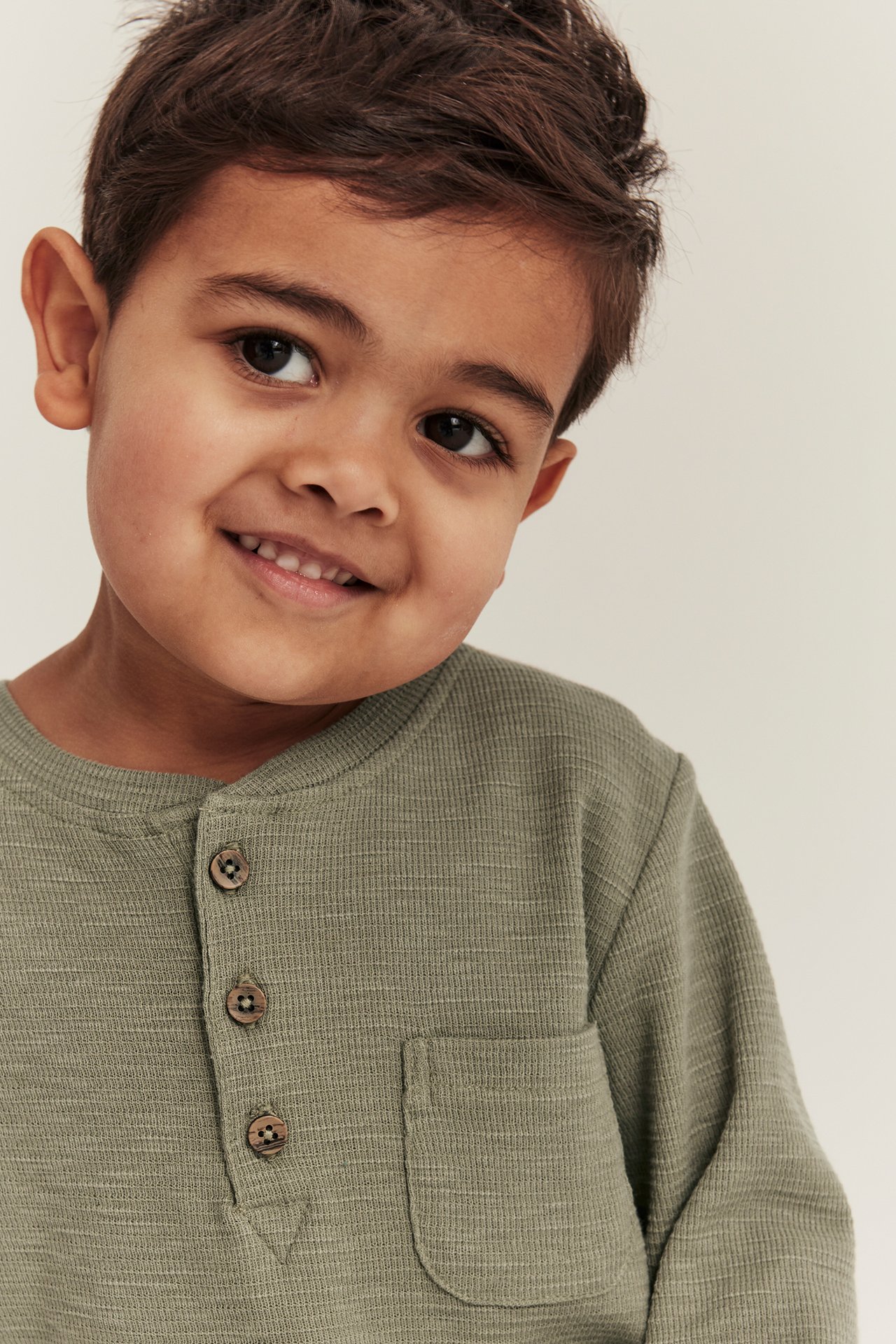 Close-up of a smiling kid in a dark green waffle-textured long-sleeved grandad shirt with buttons and pocket.
