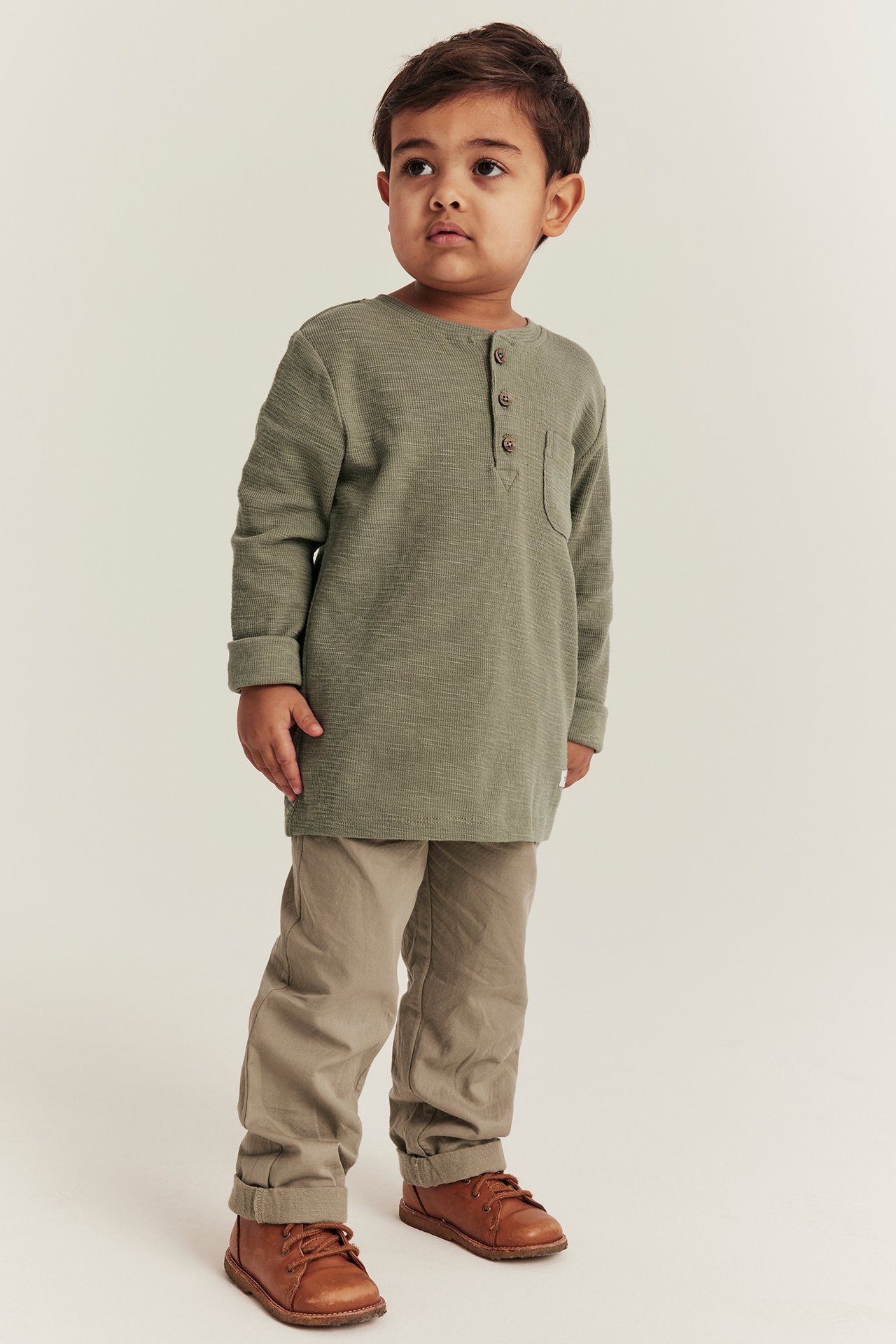 Front view of a kid in a dark green waffle-knit henley, beige pants, and brown lace-up shoes.