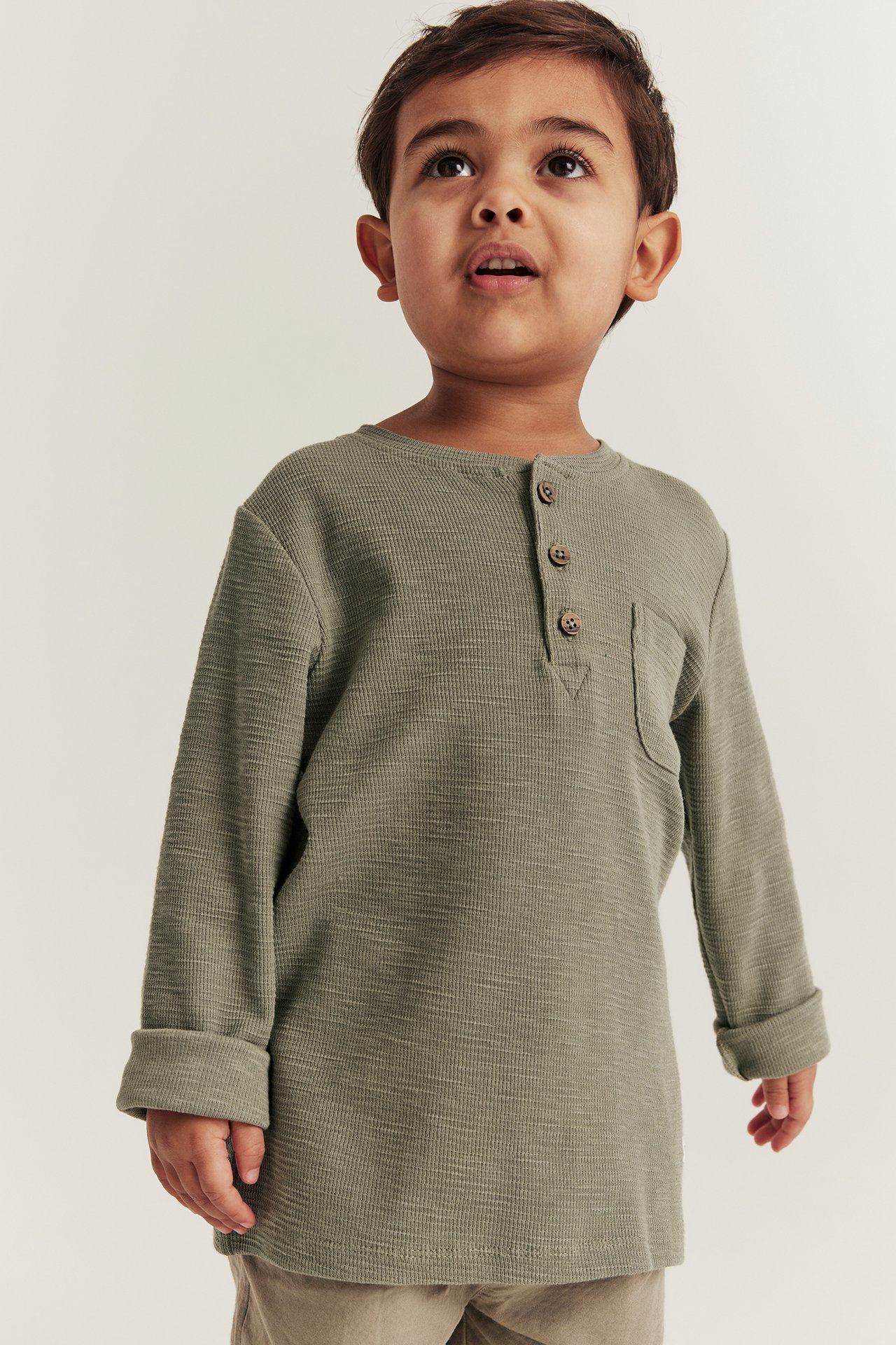 Front view of a kid in a dark green waffle henley shirt with brown buttons and beige pants.