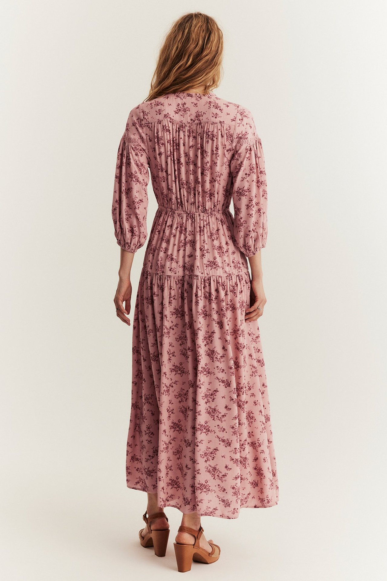 Rückenansicht einer Frau in langem rosa geblümtem Kreppviskosekleid mit Puffärmeln und braunen Sandaletten.