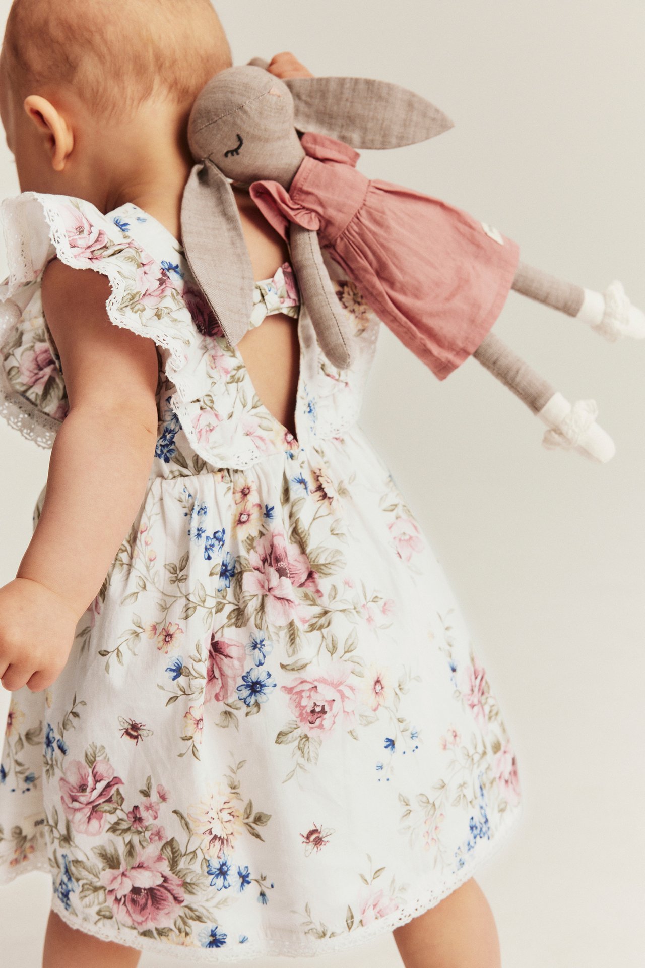 Back view of a baby in an off-white floral cotton dress with ruffles, holding a pink-dressed bunny doll.