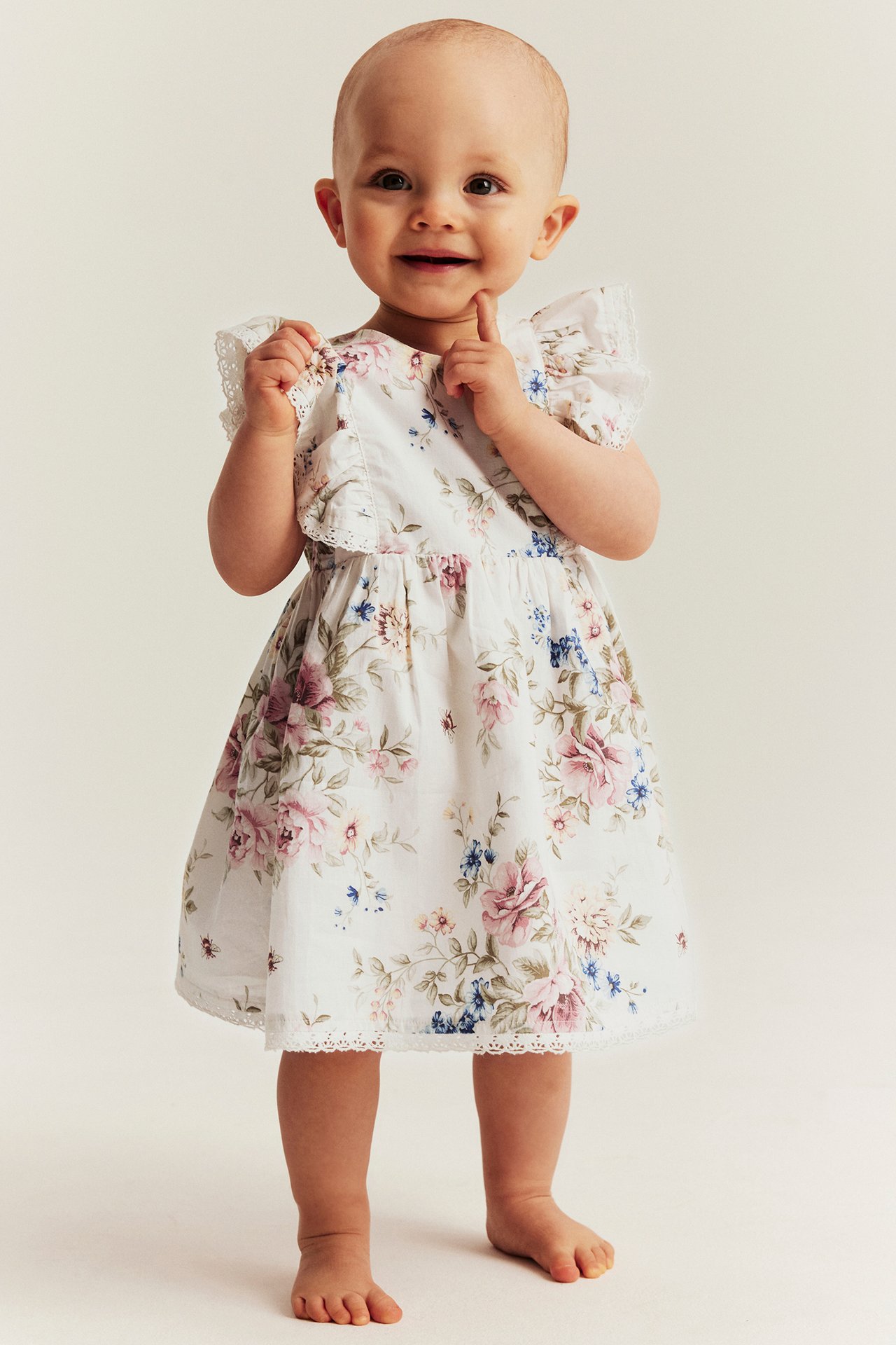 Front view of a smiling baby in an off-white floral cotton dress with ruffled, lace-trimmed sleeves.