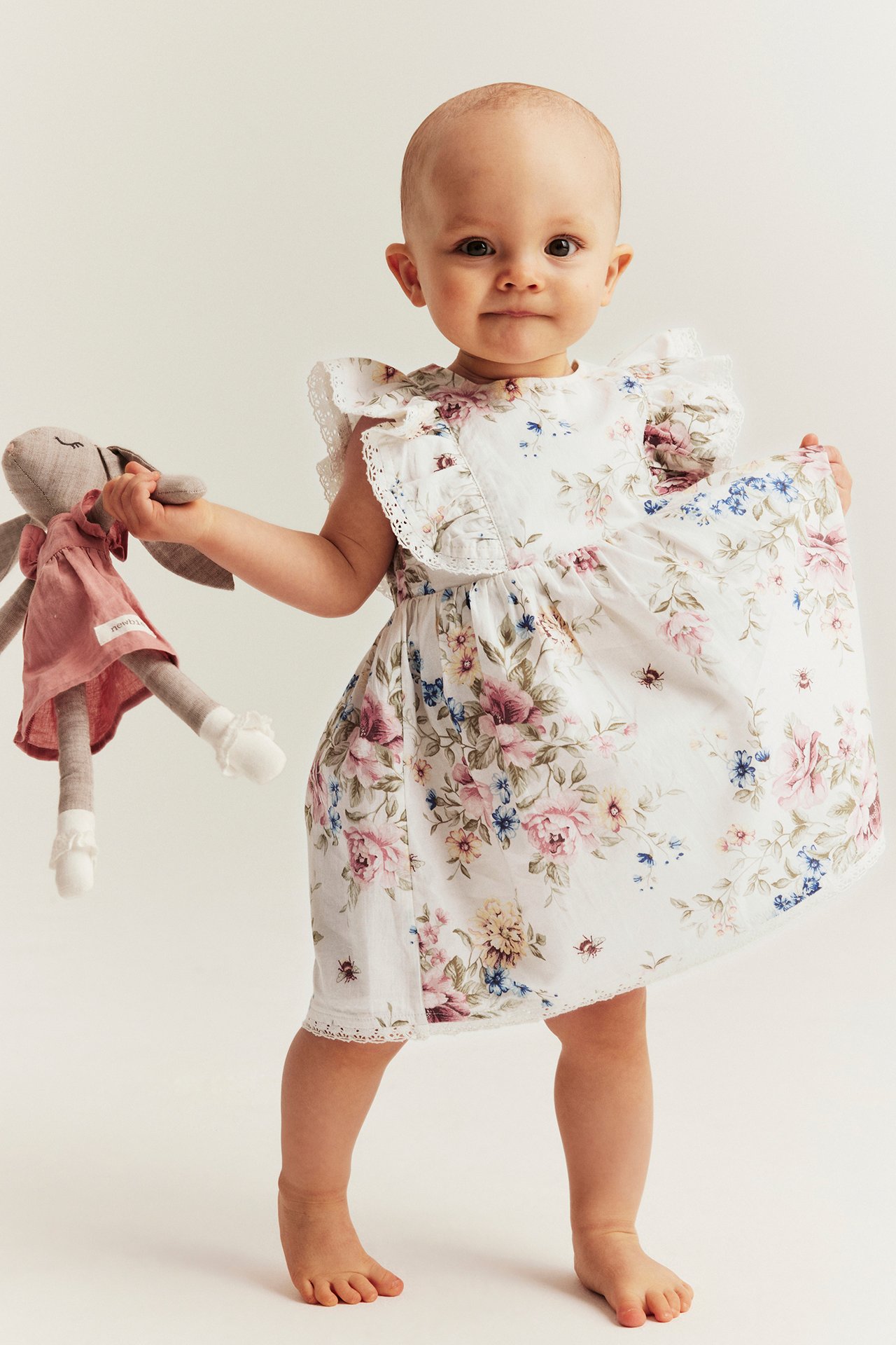 Front view: baby in off-white floral cotton dress with ruffled lace sleeves, holding a grey bunny toy.