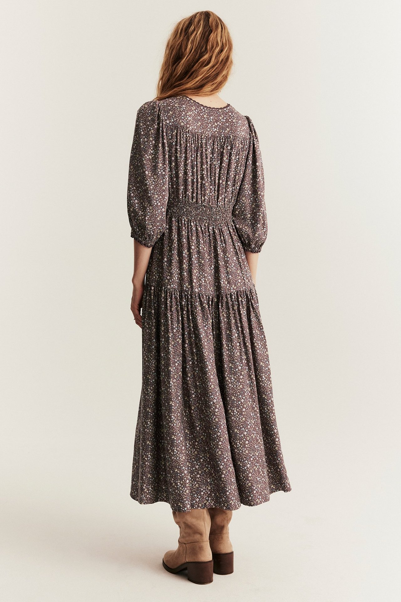 Rückenansicht einer Frau in langem lila Blumen-Viskosekleid mit gesmoktem Rücken und hellbraunen Stiefeln.