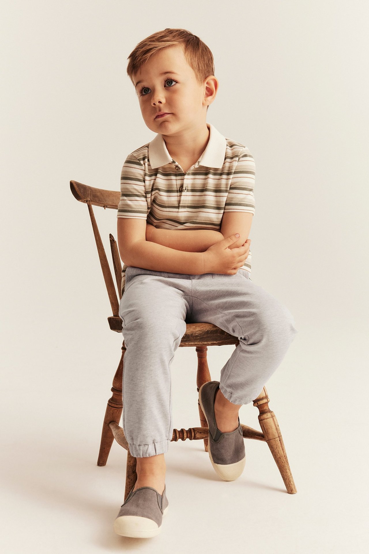 Vorderansicht eines Kindes in hellgrauen Baumwoll-Jogginghosen mit Bündchen, einem gestreiften Poloshirt und dunkelgrauen Slip-on-Schuhen.