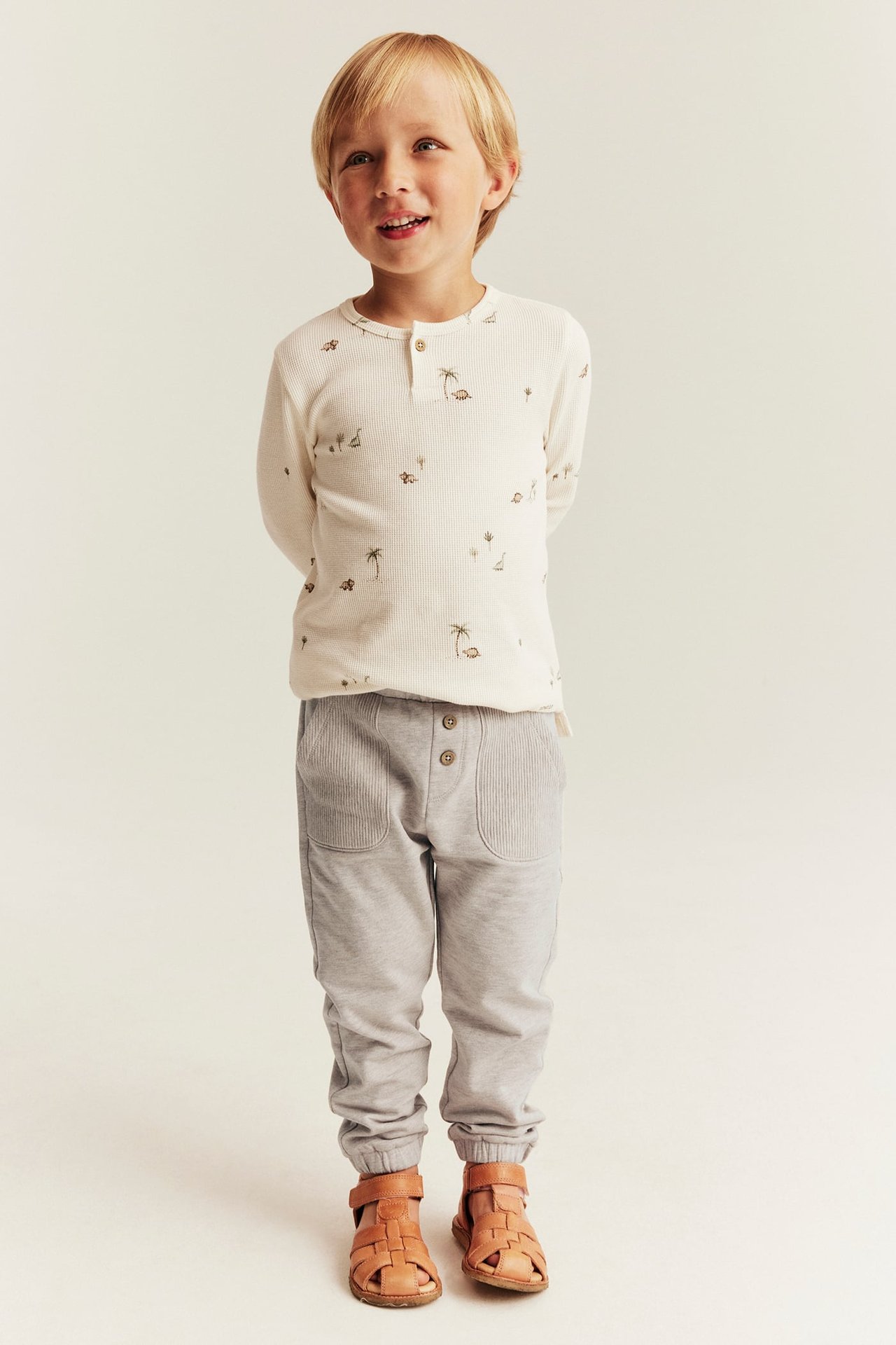 Vorderansicht Kind in cremefarbenem geripptem Henley mit Print, grauer Jogginghose, braunen Sandalen.