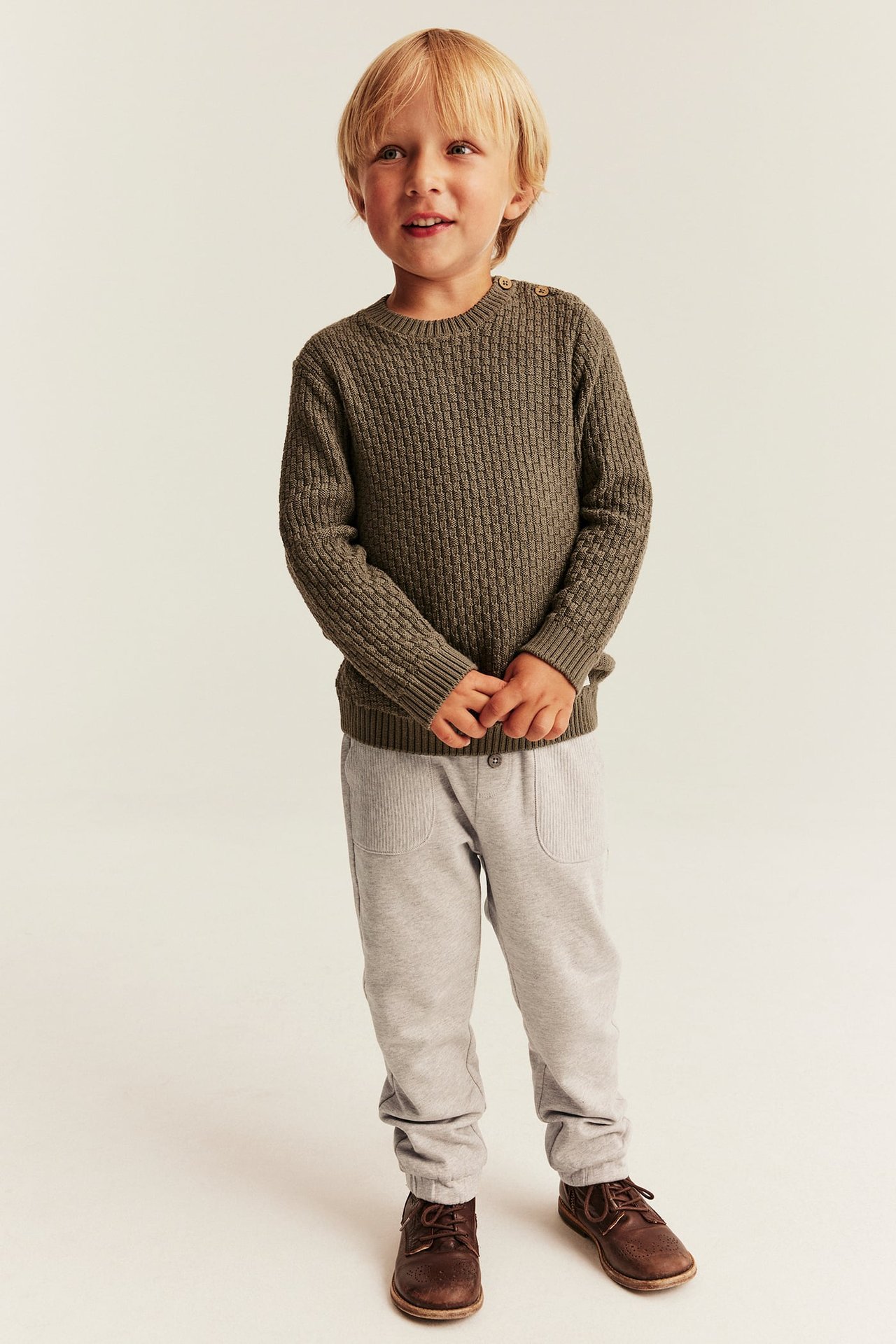Vorderansicht eines Kindes in olivgrünem Rippstrickpullover, hellgrauer Cordhose und braunen Schnürschuhen.