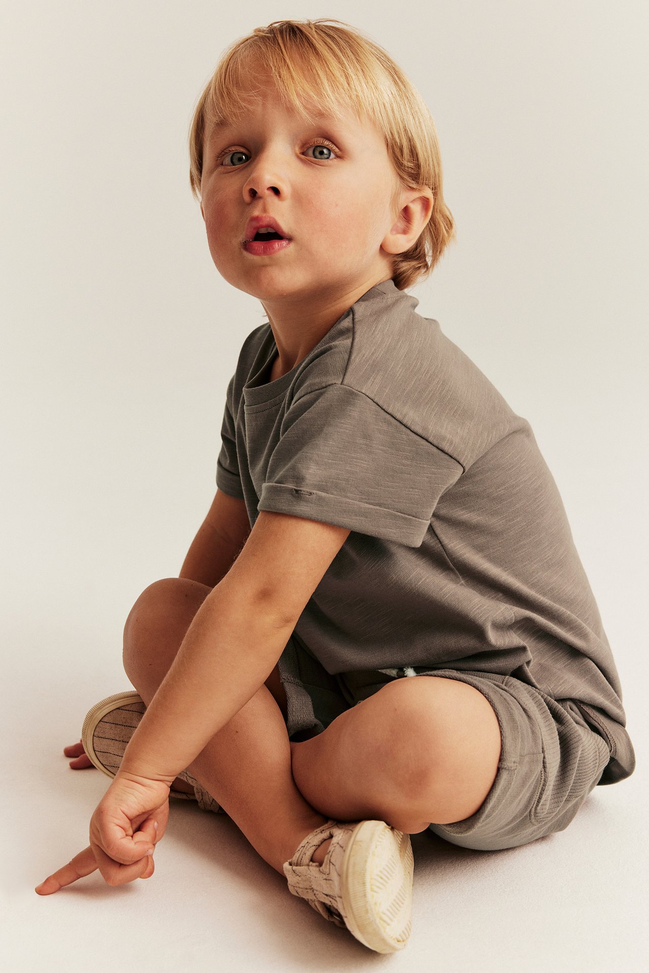 Kind in braunem Slub-Jersey-T-Shirt mit hochgekrempelten Ärmeln, passenden Ripp-Shorts und hellen Schuhen.