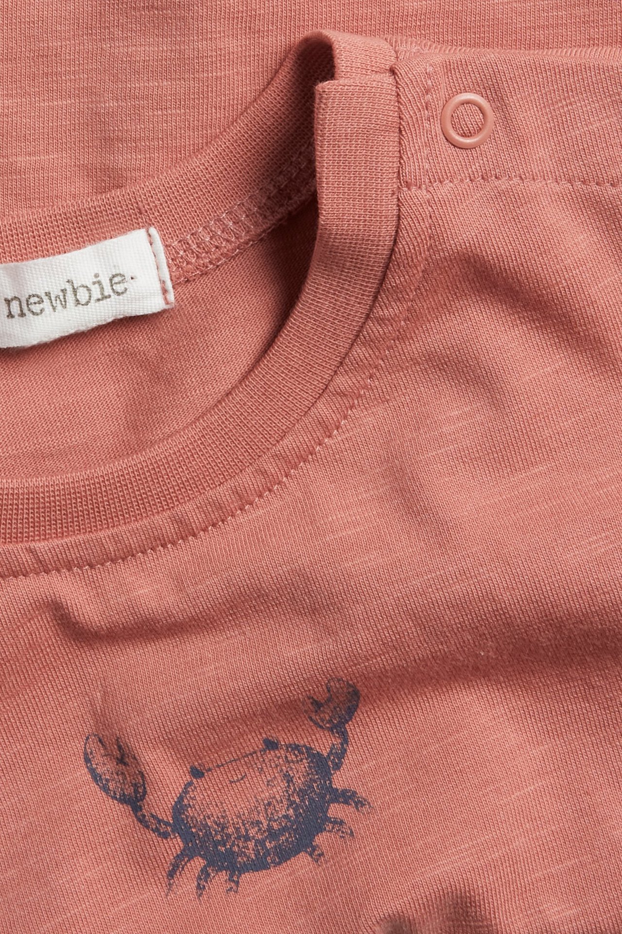 Nahaufnahme eines rosa Baumwoll-Slub-T-Shirts mit dunklem Krabbendruck und Schulter-Druckknopf.