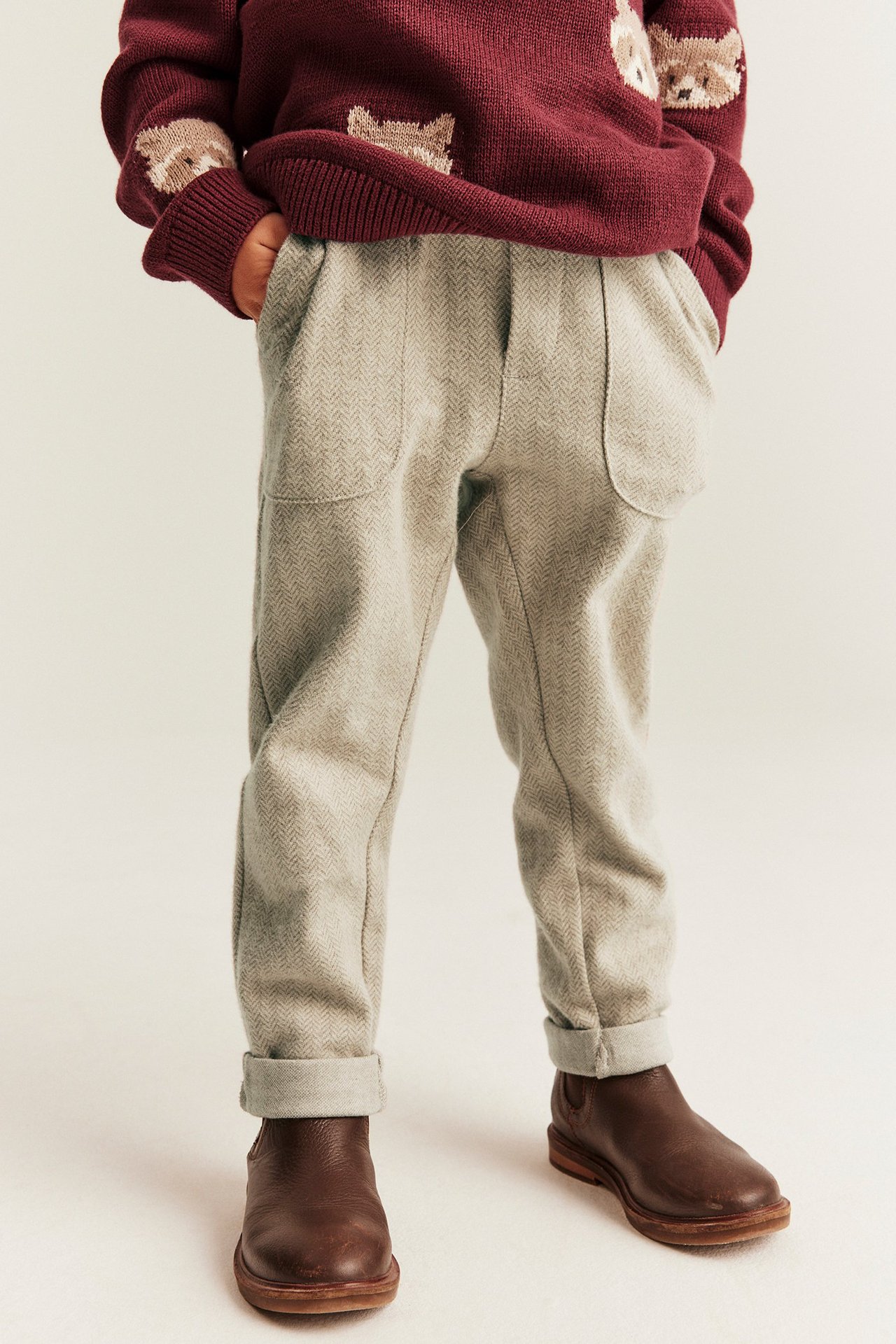 Vorderansicht: Kind trägt beige melierte Fischgrät-Hose mit umgekrempelten Säumen, roten Strickpullover und braune Stiefel.