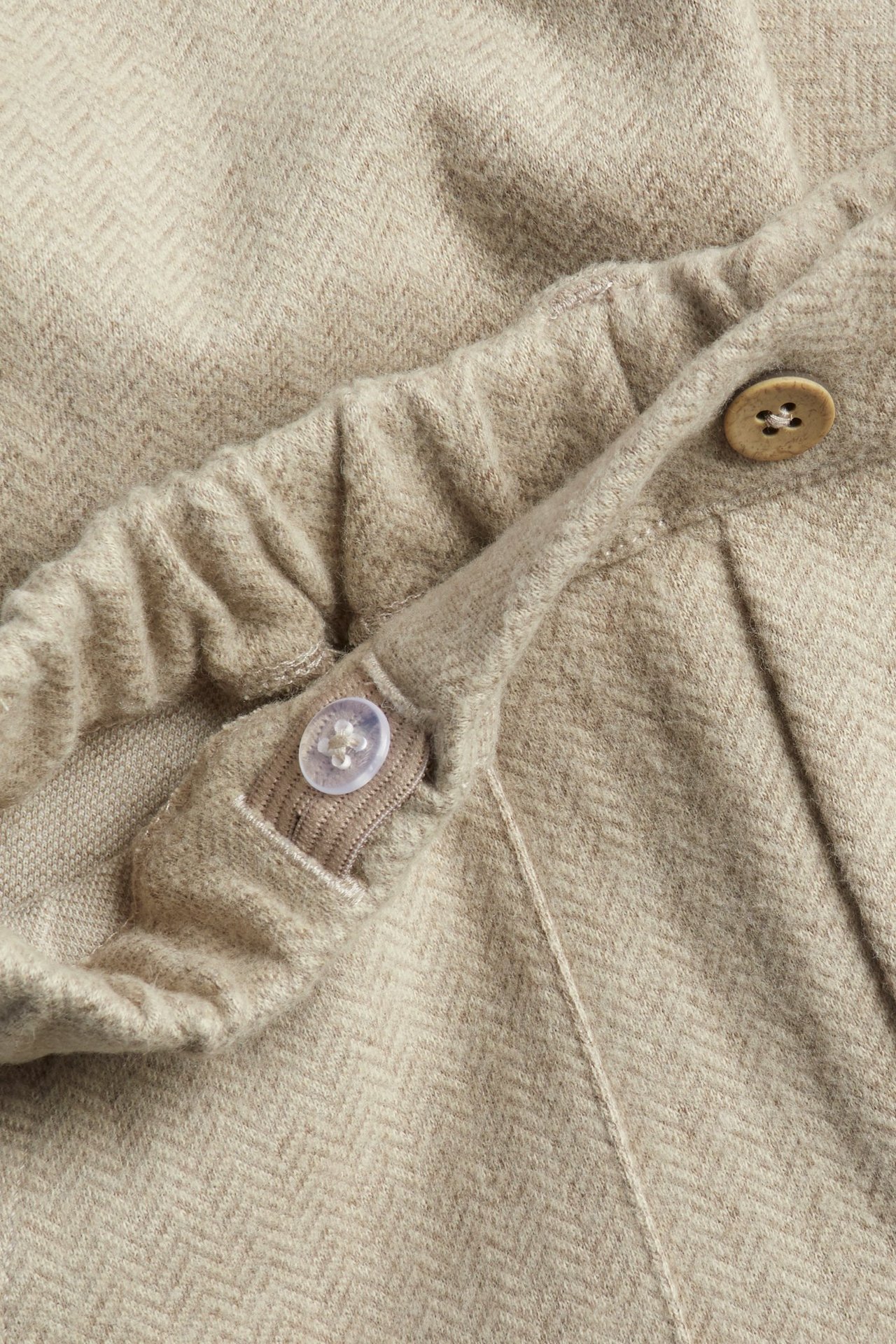Nahaufnahme beigefarbener Kinderhose mit Fischgrätmuster, elastischem Bund und dekorativen Knöpfen.