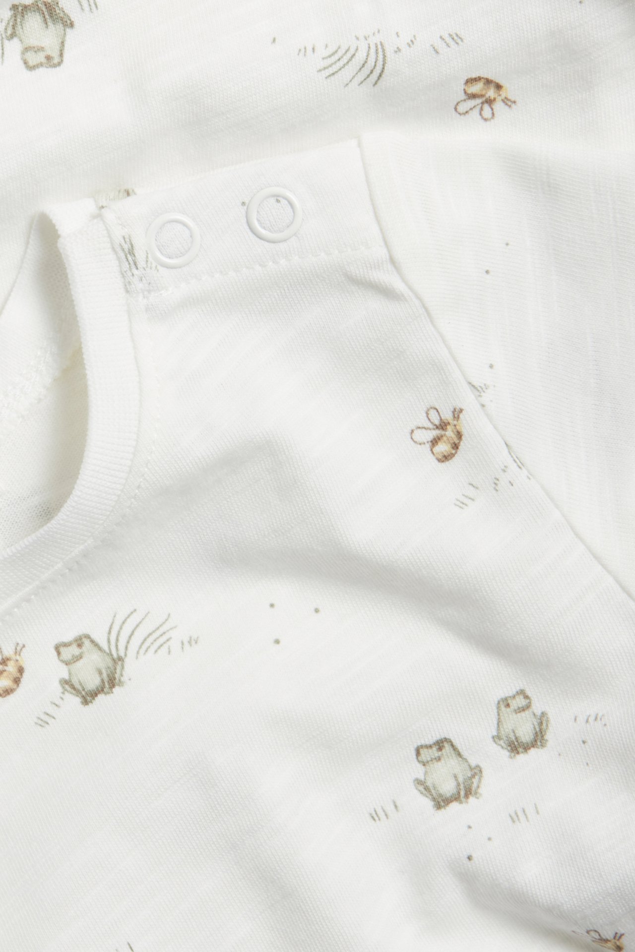 Close-up of an off-white baby short-sleeved cotton bodysuit with frog and bee pattern, featuring shoulder snap buttons.