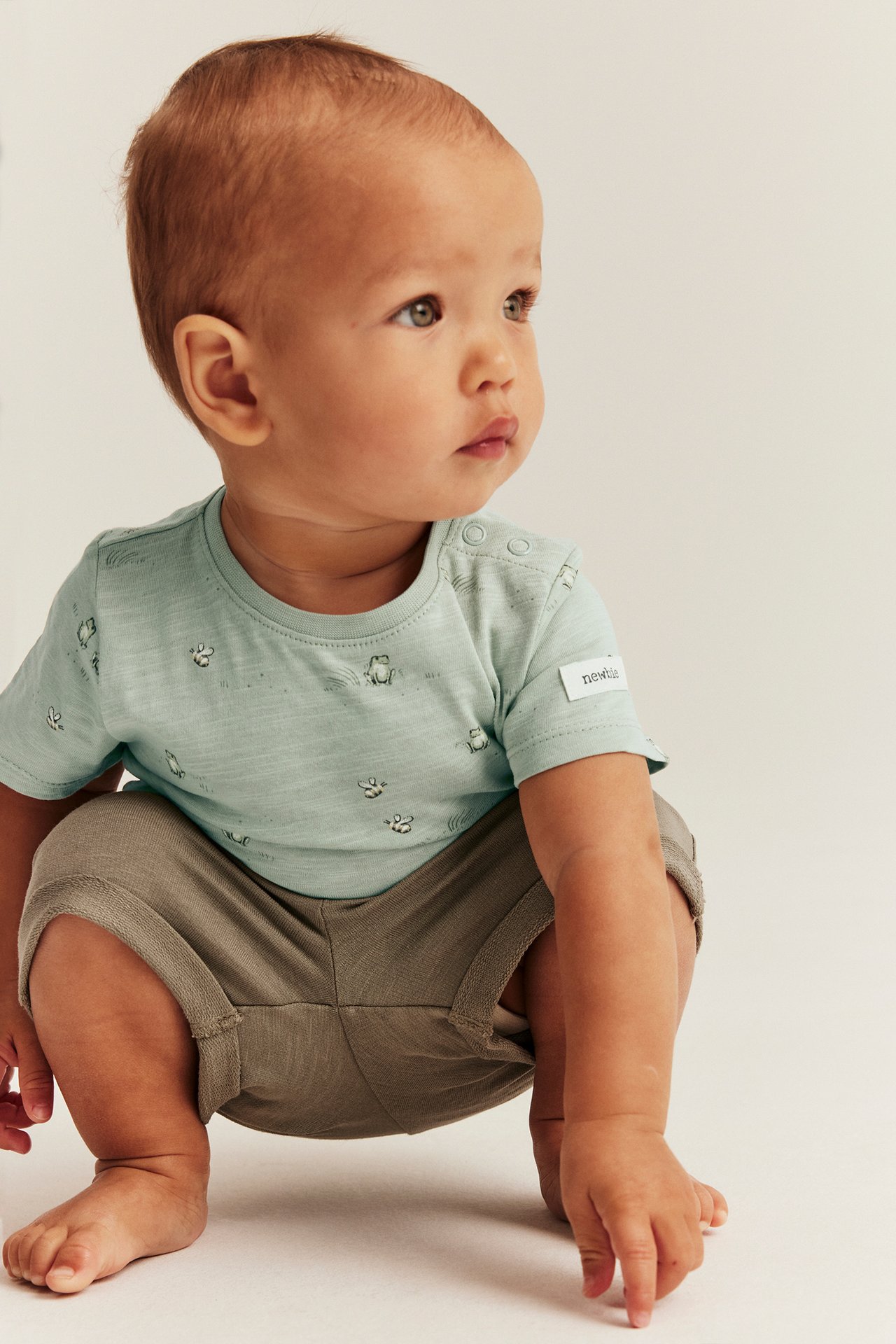 Seitenansicht eines Babys in hellgrünem kurzärmeligem Froschmuster-Baumwollbody und khakifarbenen Shorts.