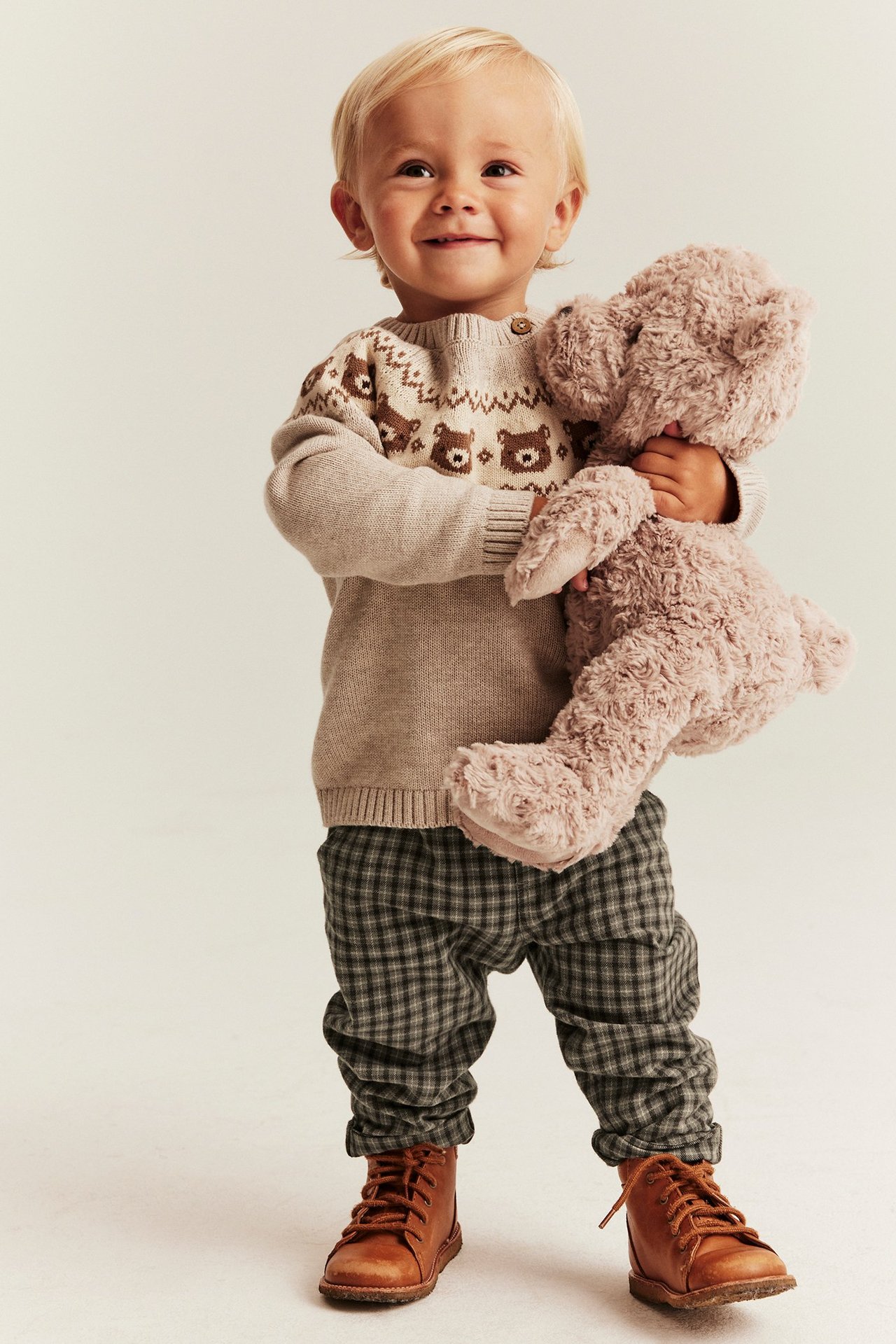 Vorderansicht: Baby in brauner karierter Webhose, beigem Bärenpullover und braunen Stiefeln, hält einen Teddybären.