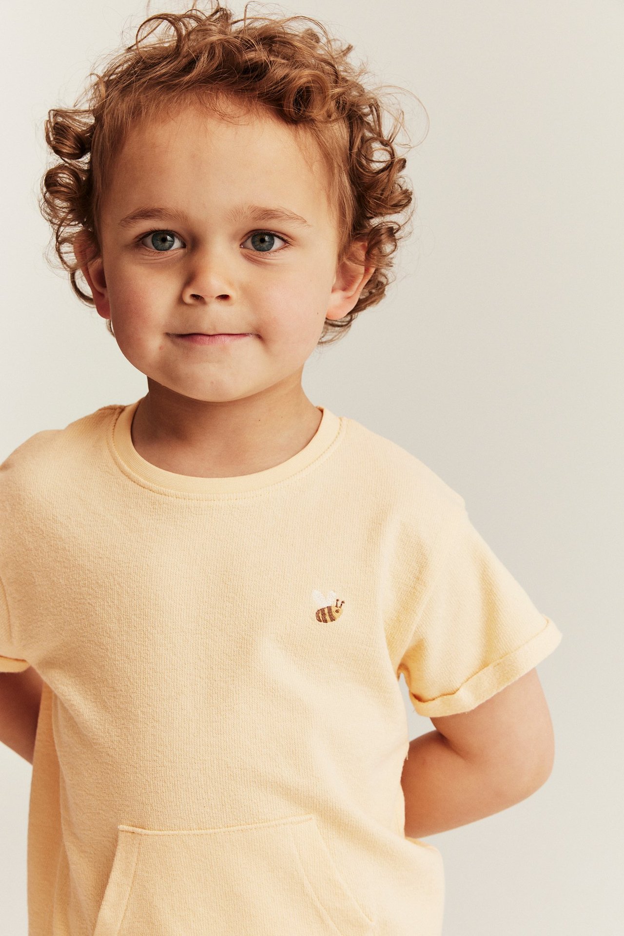 Vorderansicht eines Kindes in einem gelben Kurzarm-T-Shirt mit Bienenstickerei und Vordertasche.