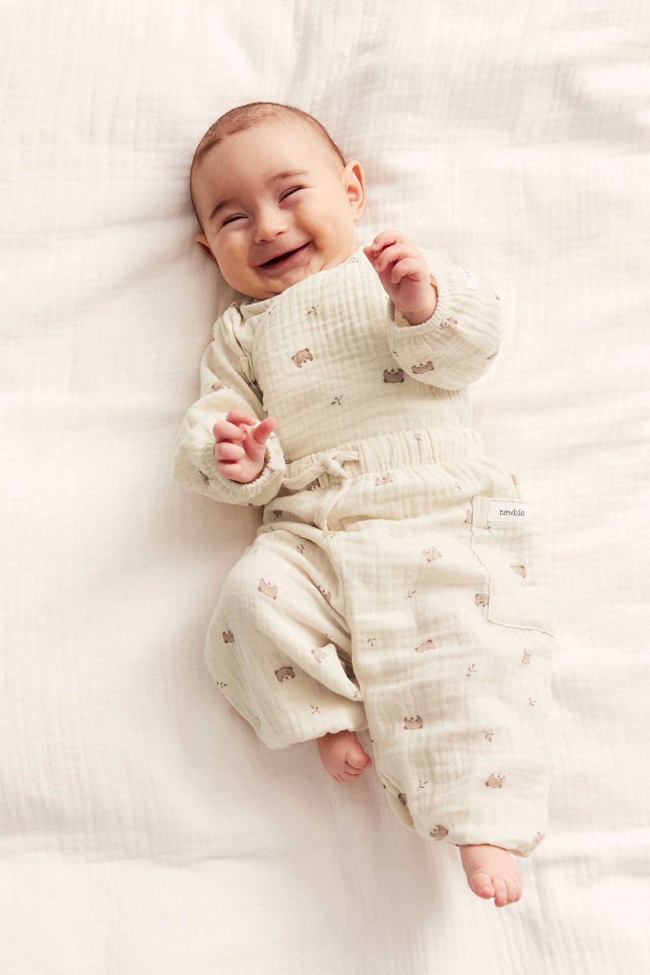 Top-down view of a smiling baby wearing light beige cotton muslin animal-patterned pants and a matching long-sleeved body.