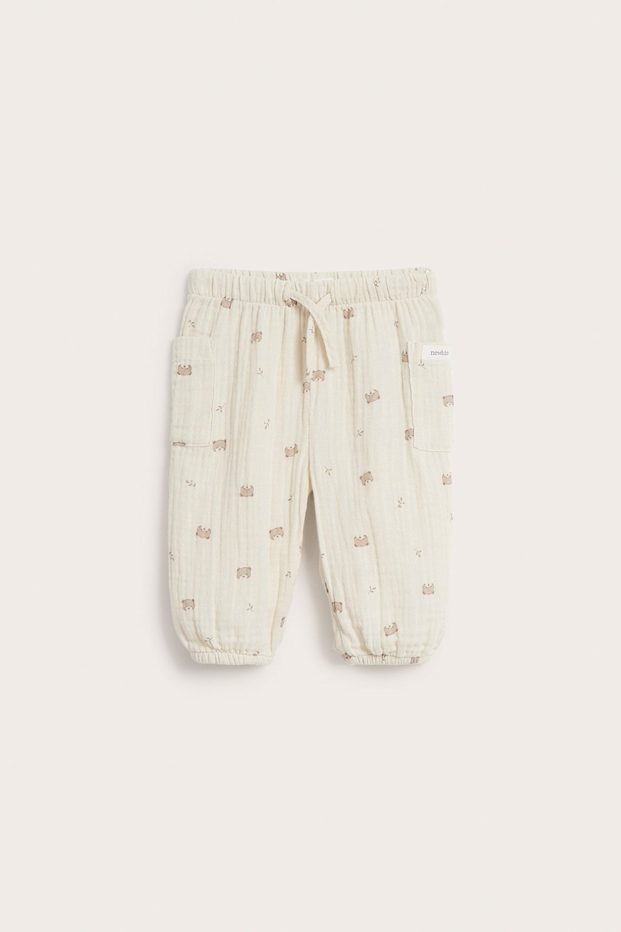Front view: Light beige cotton muslin baby pants with brown bear pattern, elastic waist, drawstring, two front pockets.