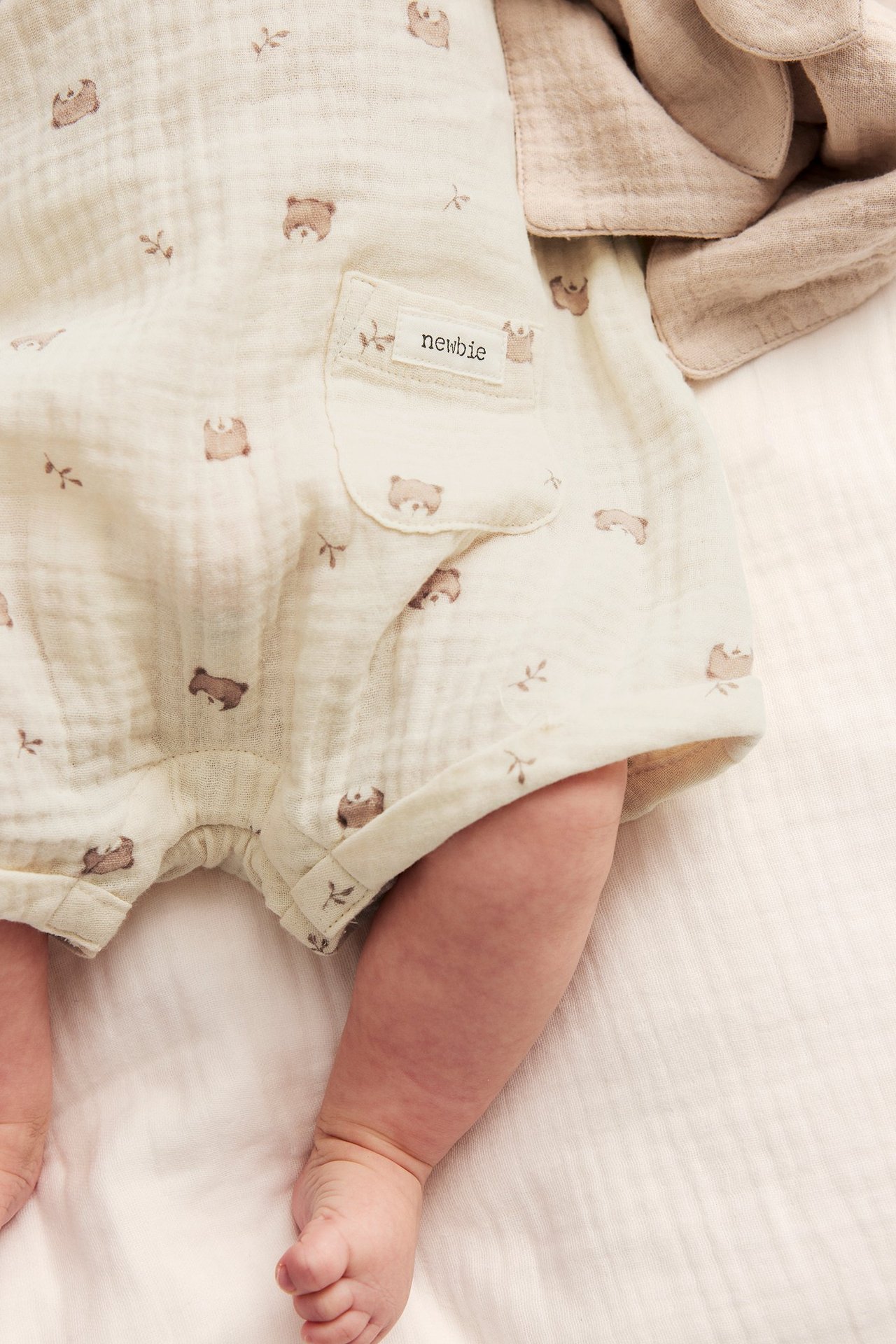 Close-up of a baby wearing a light beige cotton playsuit with a brown teddy bear pattern and front pocket.