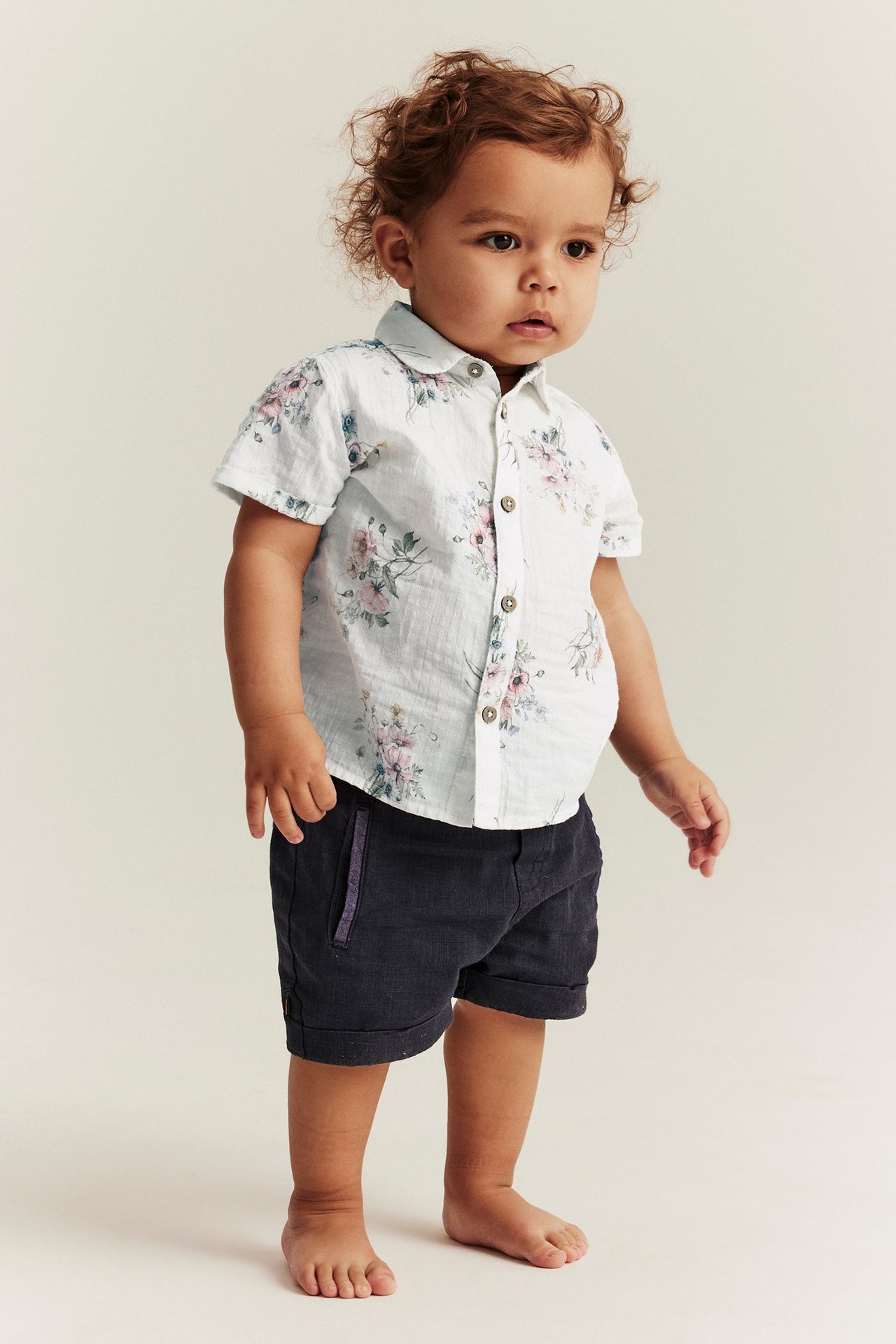 Vorderansicht eines Babys in einem cremefarbenen geblümten Kurzarmhemd mit Knöpfen und dunklen Shorts.