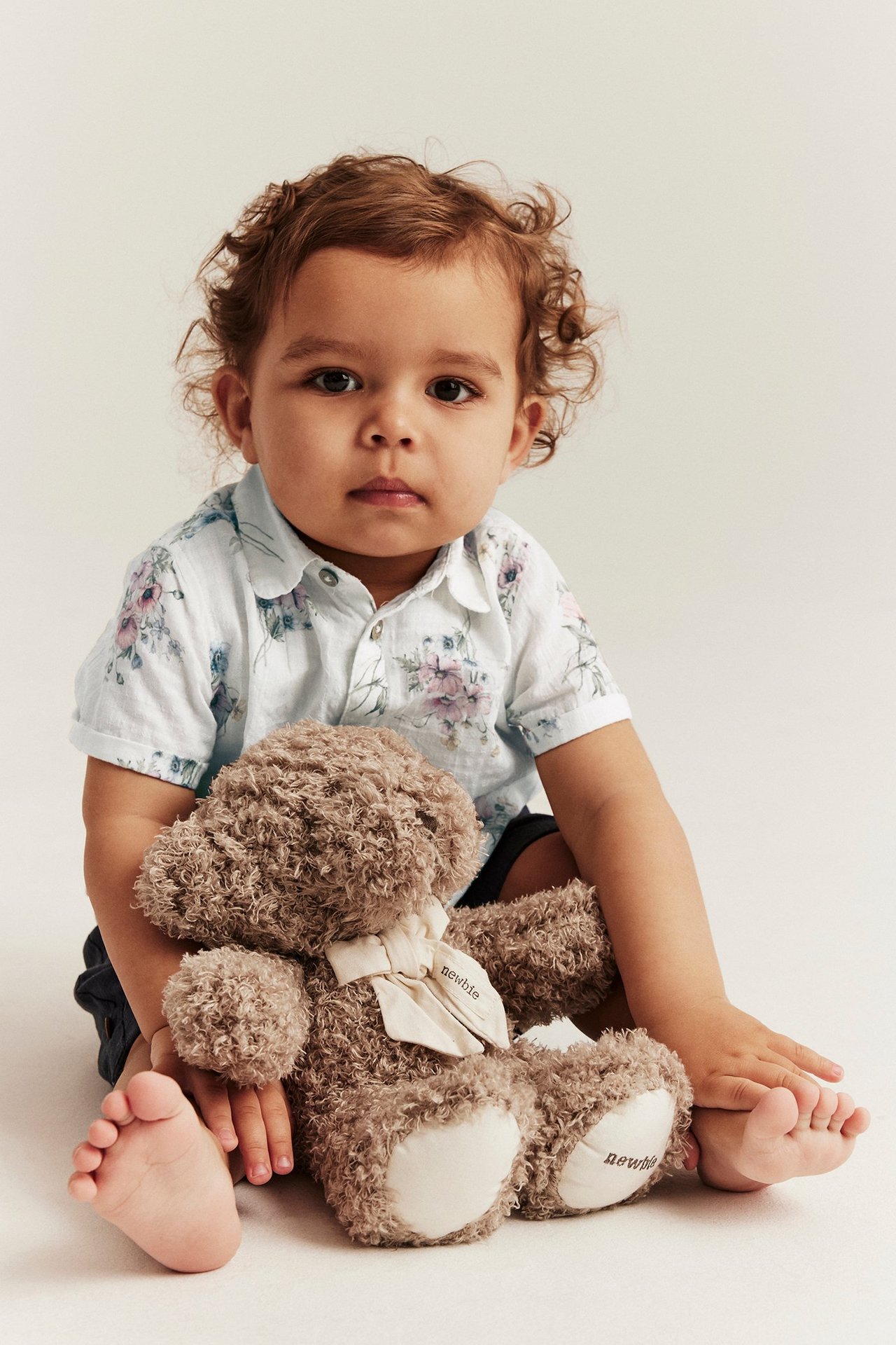 Vorderansicht: Baby in einem cremefarbenen, geblümten kurzärmeligen Baumwollhemd, dunklen Shorts, hält einen braunen Teddybär.