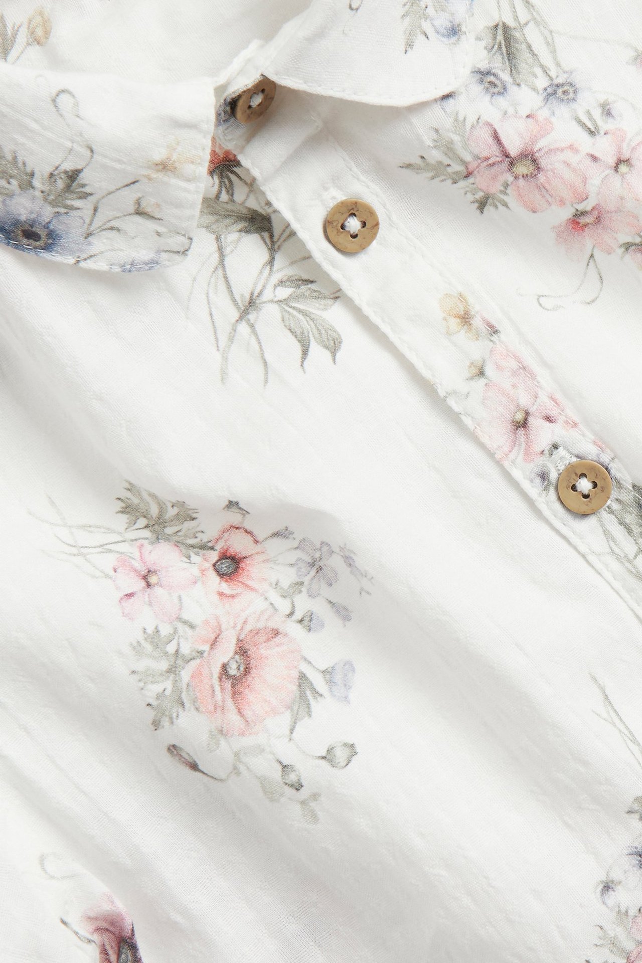 Nahaufnahme eines cremefarbenen kurzärmeligen Baumwollhemdes mit Blumenmuster und Holzknöpfen.