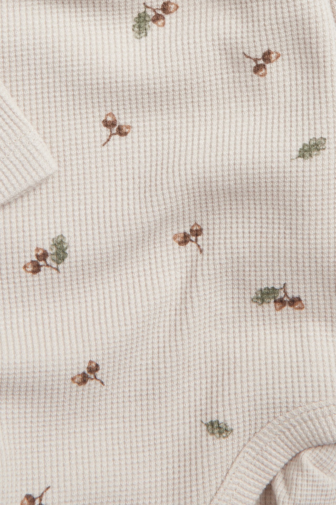 Close-up of a beige waffle-knit long-sleeved baby bodysuit with a brown acorn and green leaf pattern.