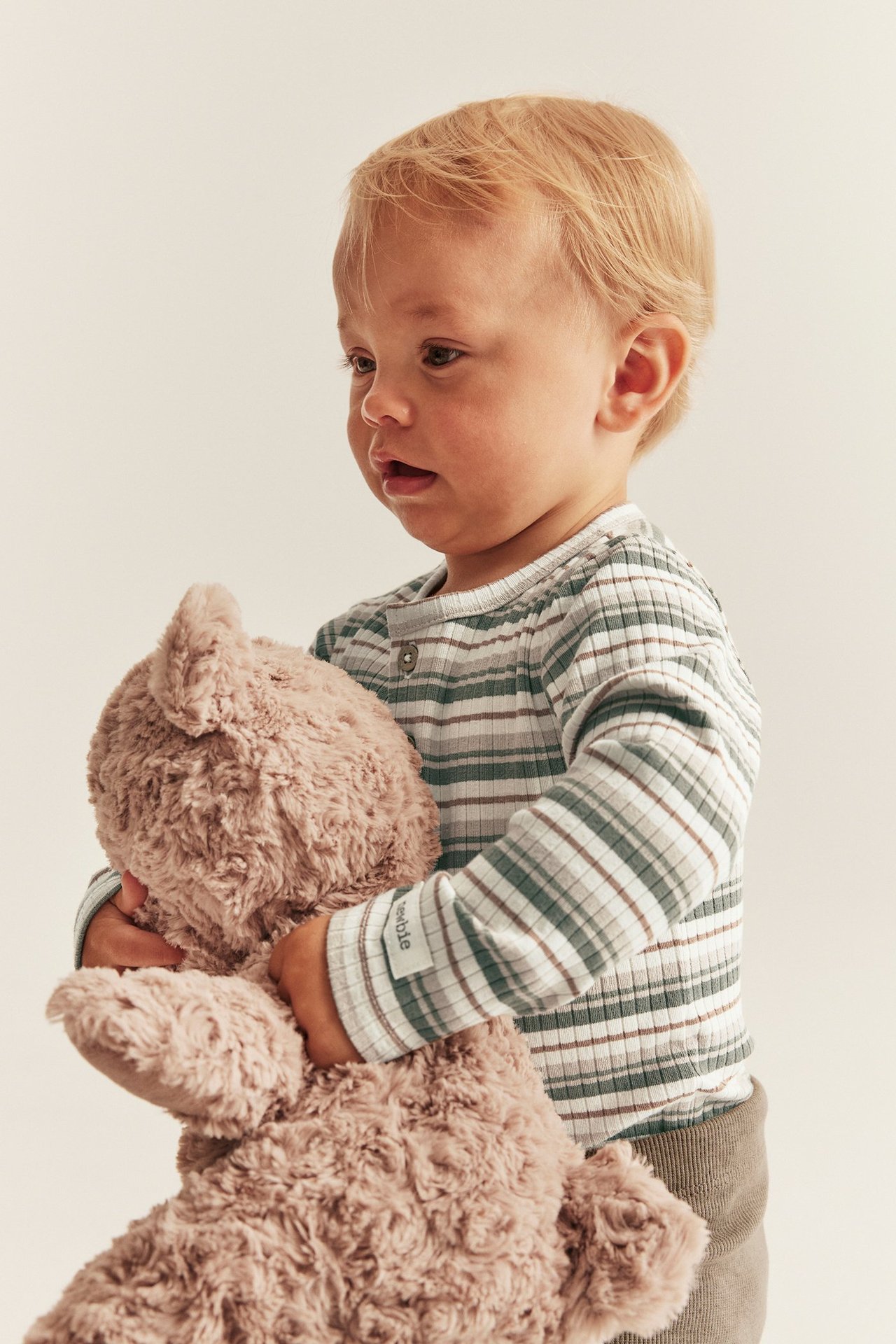 Nahaufnahme eines Babys in einem gerippten, gestreiften Langarm-Body in Off-White, das einen braunen Teddy hält.