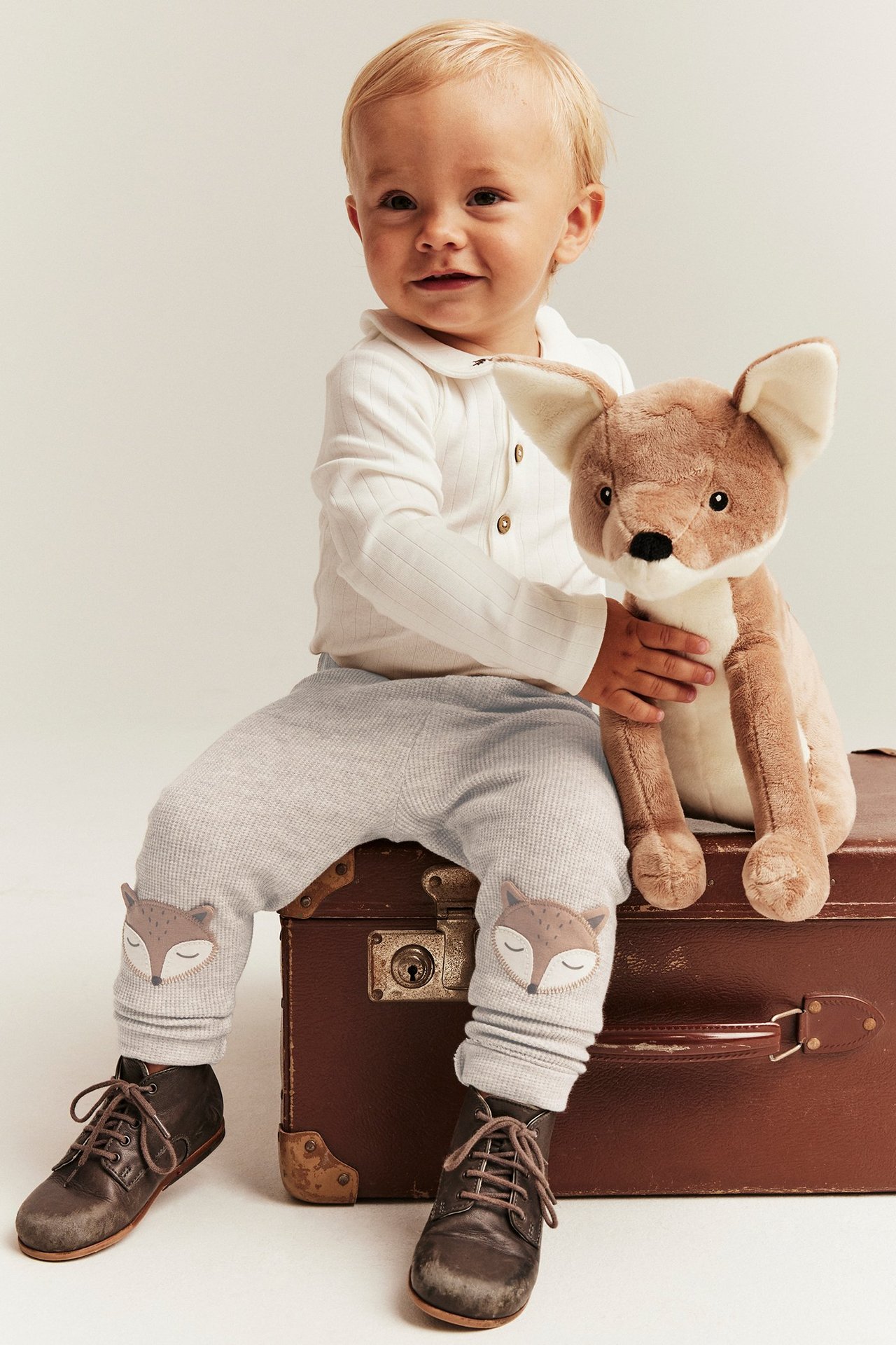 Baby in hellgrauen Waffel-Leggings mit Fuchs-Knieapplikationen, weißem Ripp-Top und braunen Stiefeln.