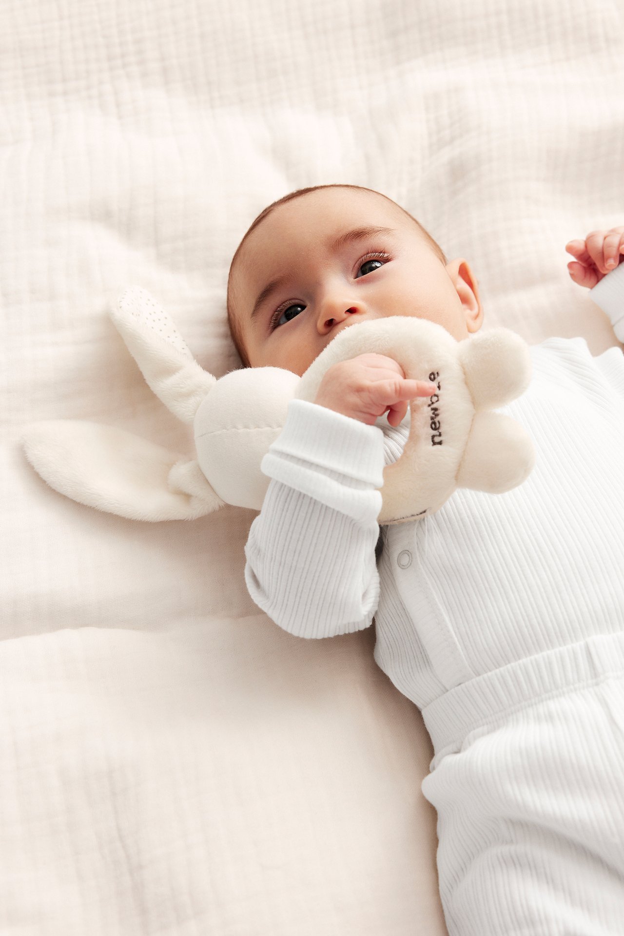 Nahaufnahme eines Babys in einem cremefarbenen gerippten Wickelbody, das eine cremefarbene Plüschhasenrassel hält.