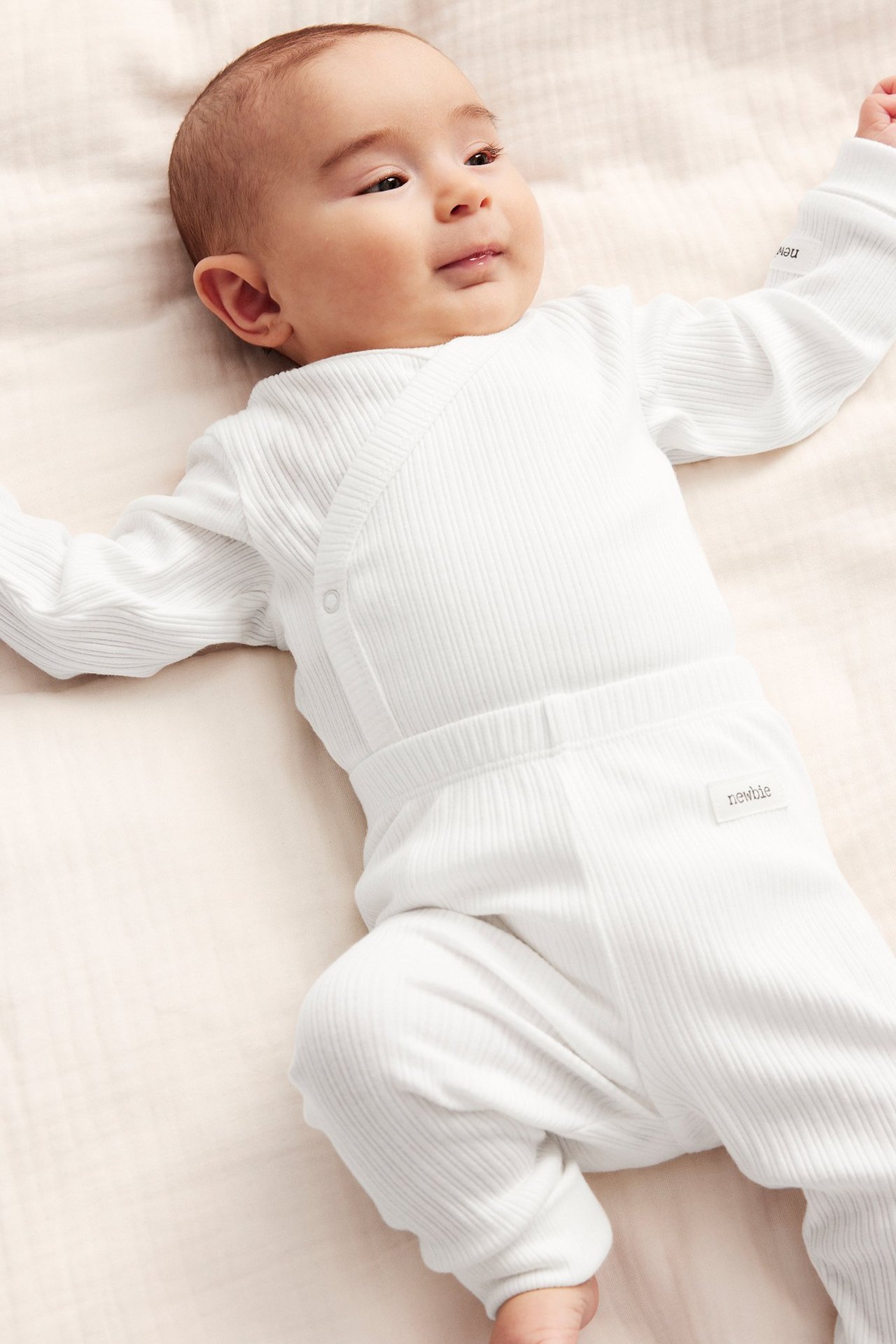 Vorderansicht eines Babys in einem gerippten, cremefarbenen Wickelbody mit Druckknöpfen und passender Hose.