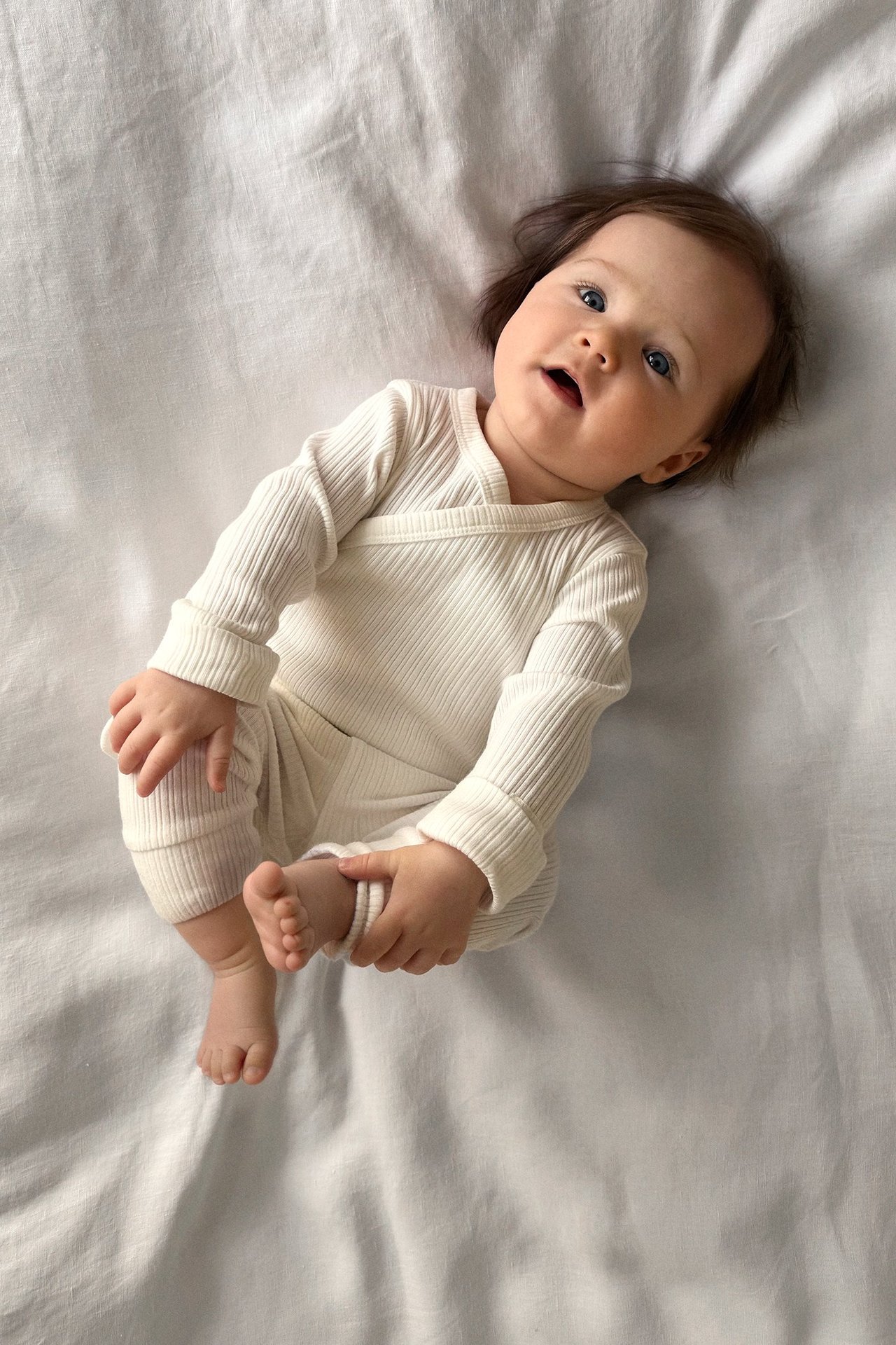 Draufsicht auf ein Baby in einem gerippten, cremeweißen Wickelbody mit langen Ärmeln und umklappbaren Bündchen.