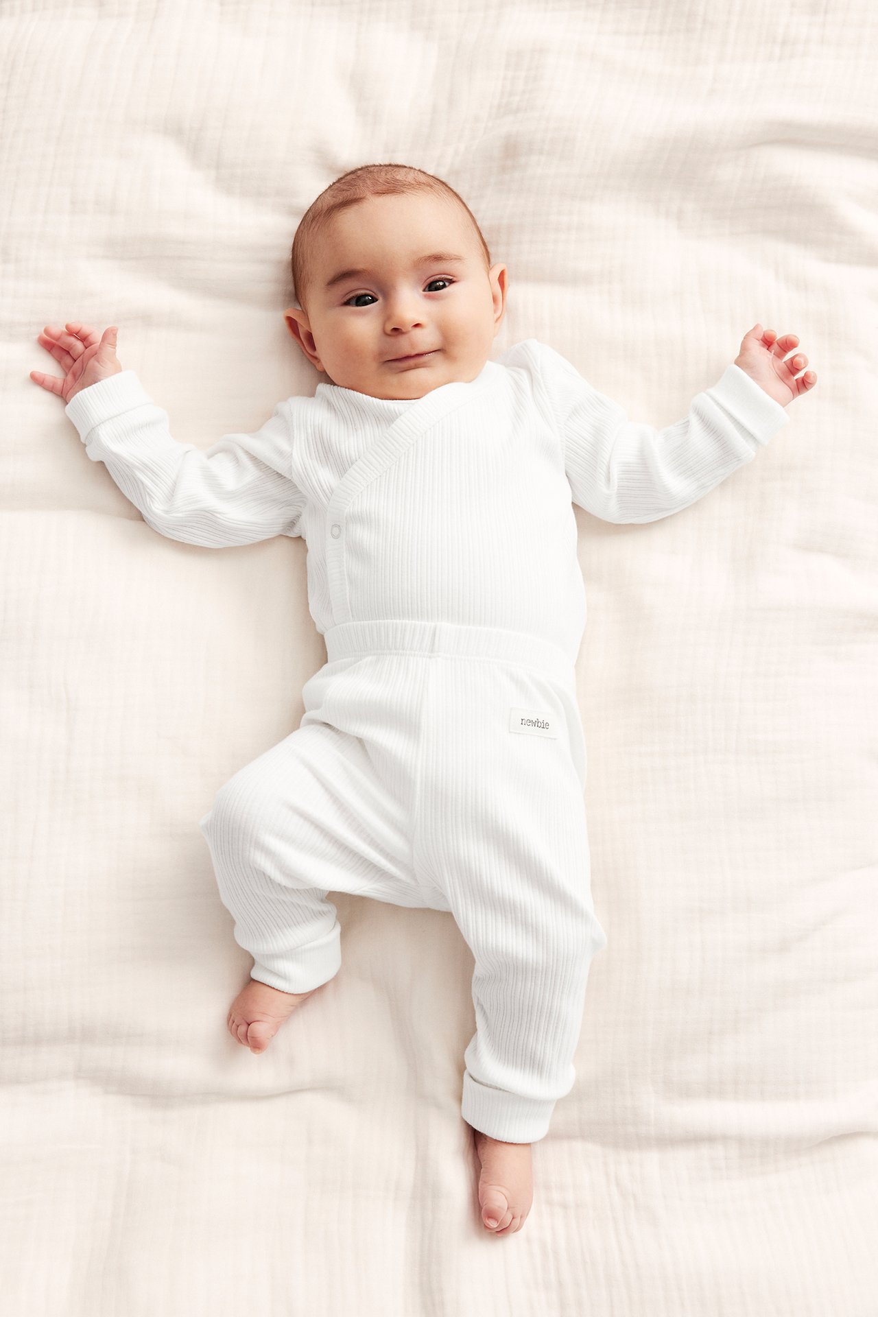 Draufsicht: Baby in cremeweißen gerippten Leggings mit umgeschlagenen Bündchen und passendem gerippten Wickeloberteil.