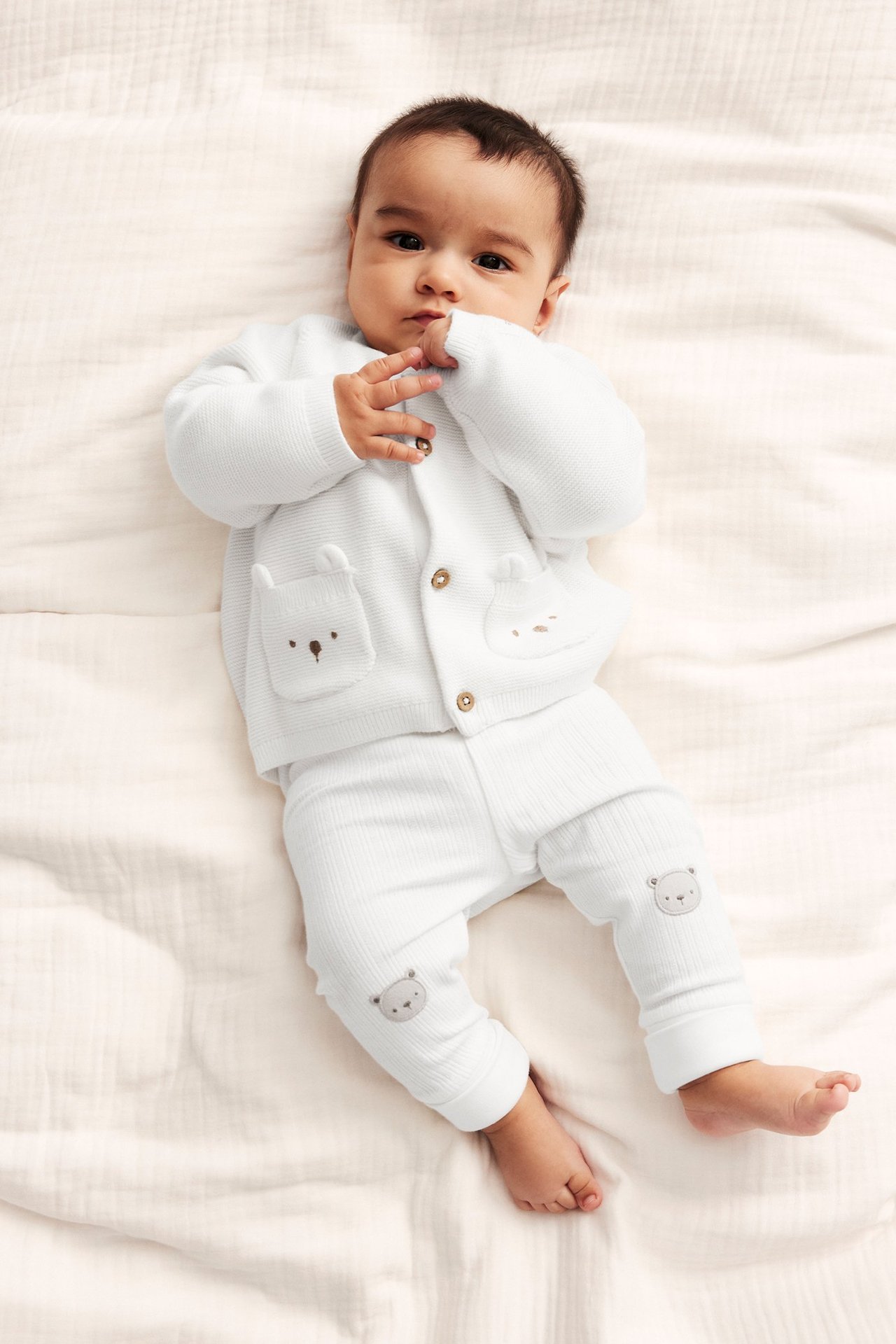 Draufsicht: Baby in cremeweißer Baumwollstrickjacke mit Bärenstickerei-Taschen und passender Ripphose.