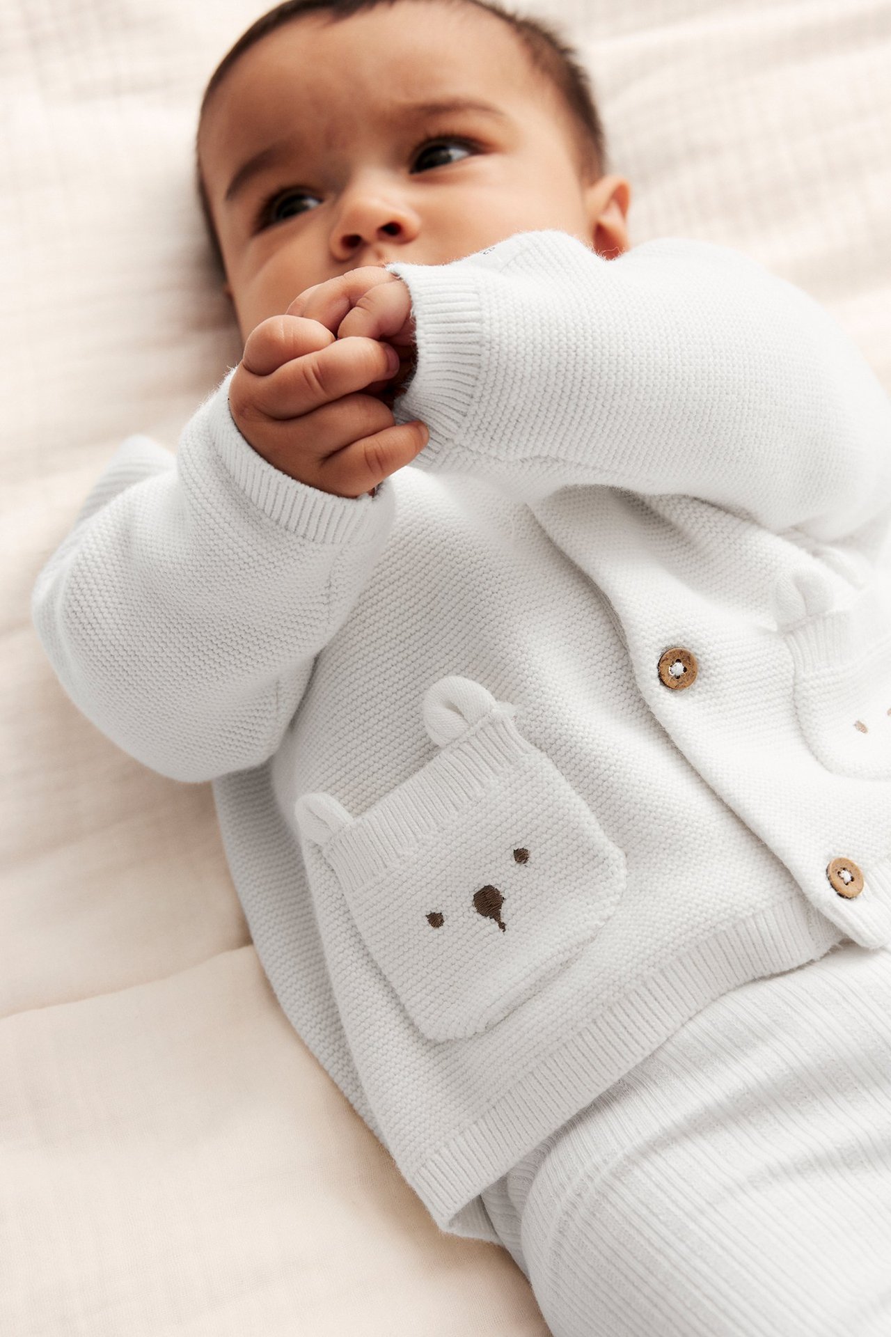 Nahaufnahme eines Babys in cremeweißer Baumwoll-Strickjacke mit Bärengesicht-Taschen und passender Hose.