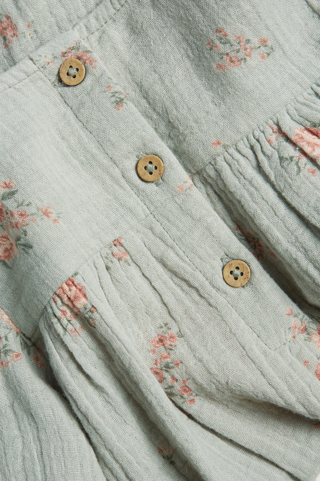 Close-up of a light green woven cotton dress with small pink floral print, gathers, and wooden buttons.