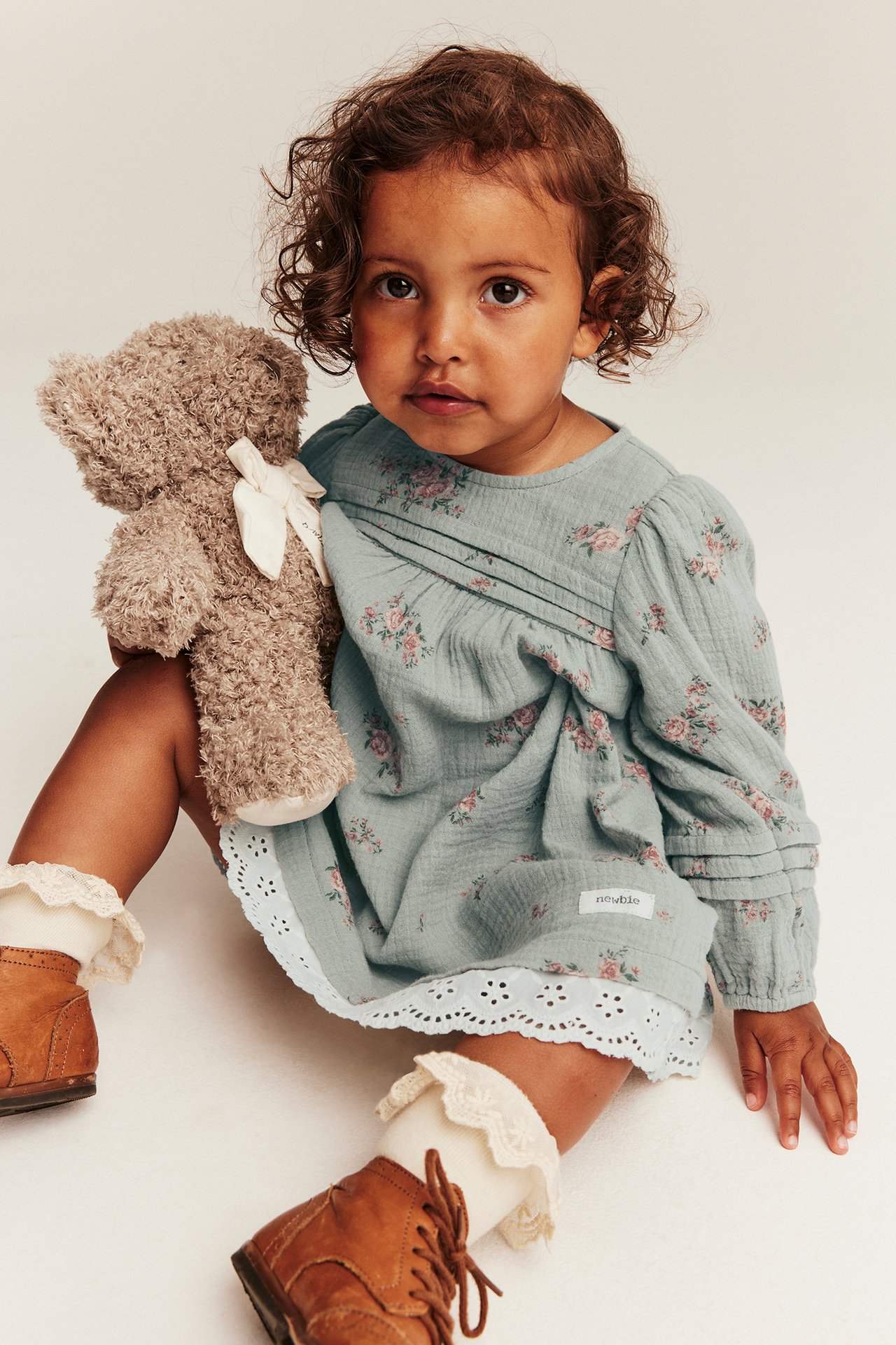 Front view of a kid wearing a light green floral woven cotton dress with pintucks and lace trim, holding a teddy bear.