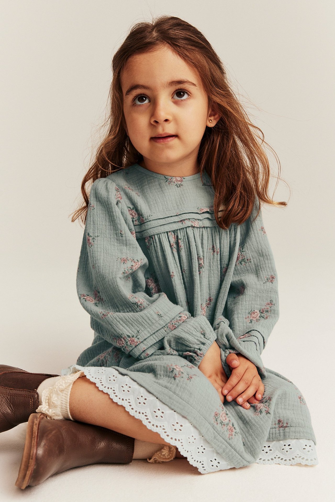 Front view of a kid in a light green floral cotton dress with pintucks, lace hem, and brown boots.
