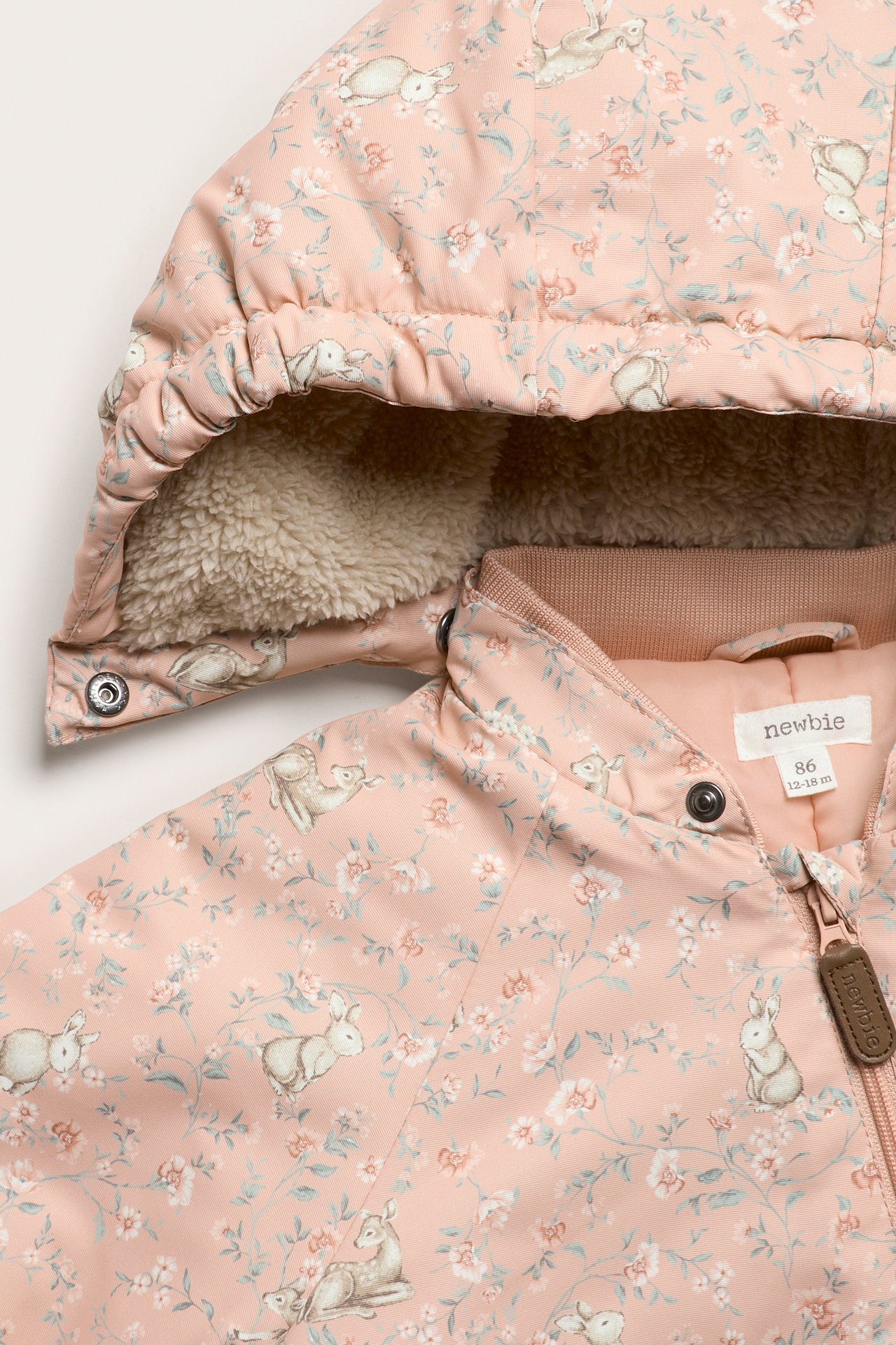 Nahaufnahme eines rosa Babyoveralls mit Blumen- und Tiermuster, flauschig gefütterter Kapuze und Reißverschluss.