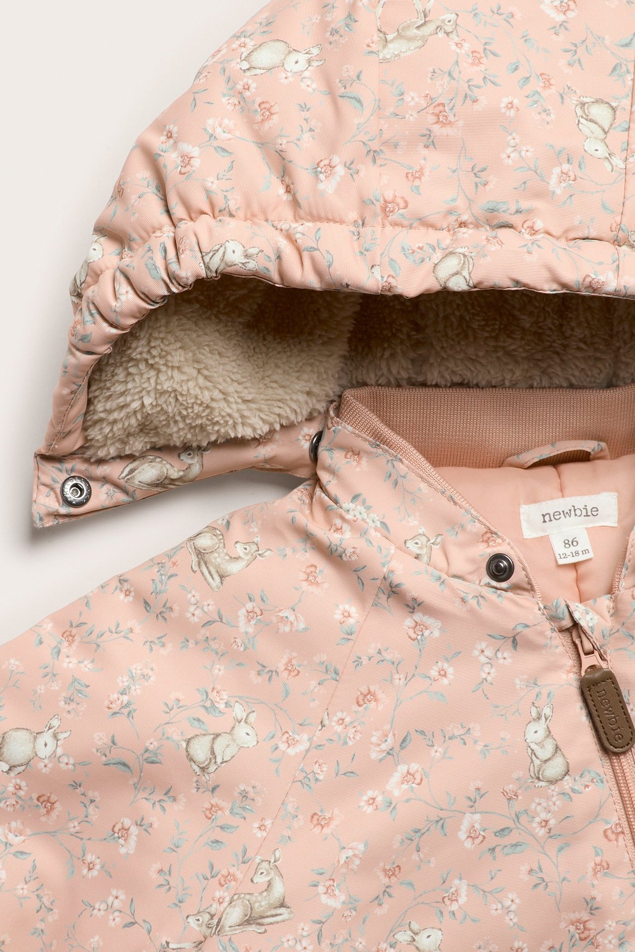 Nahaufnahme eines rosa Babyoveralls mit Blumen- und Tiermuster, flauschig gefütterter Kapuze und Reißverschluss.
