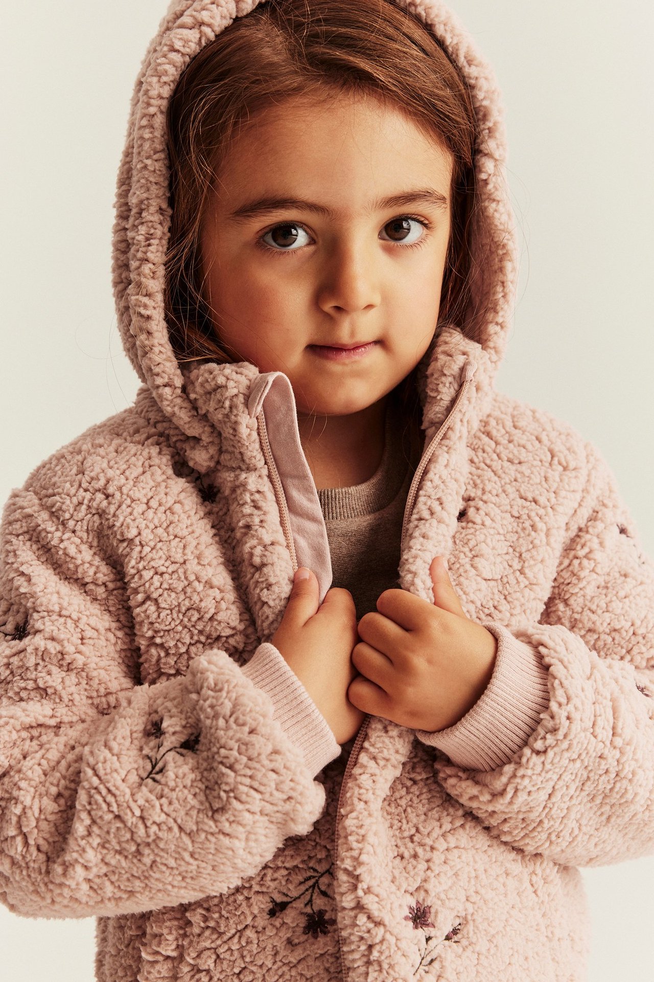 Nahaufnahme von vorne eines Kindes in einer rosa Kapuzen-Pilejacke mit Blumenstickerei und Reißverschluss.