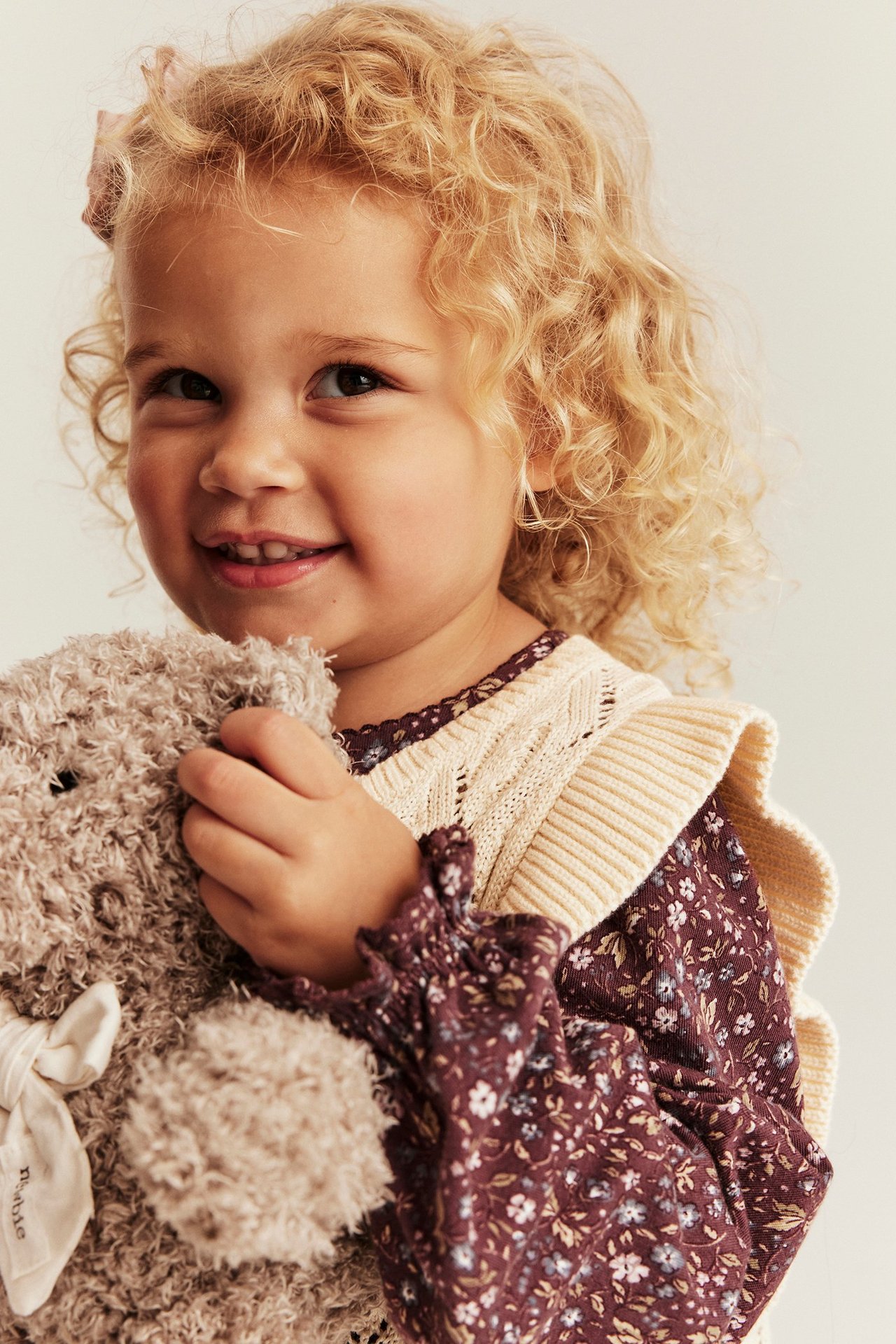 Nahaufnahme eines Kindes in einer hellbeigen gemusterten Strickweste über einem Blümchen-Oberteil, hält einen Teddy.