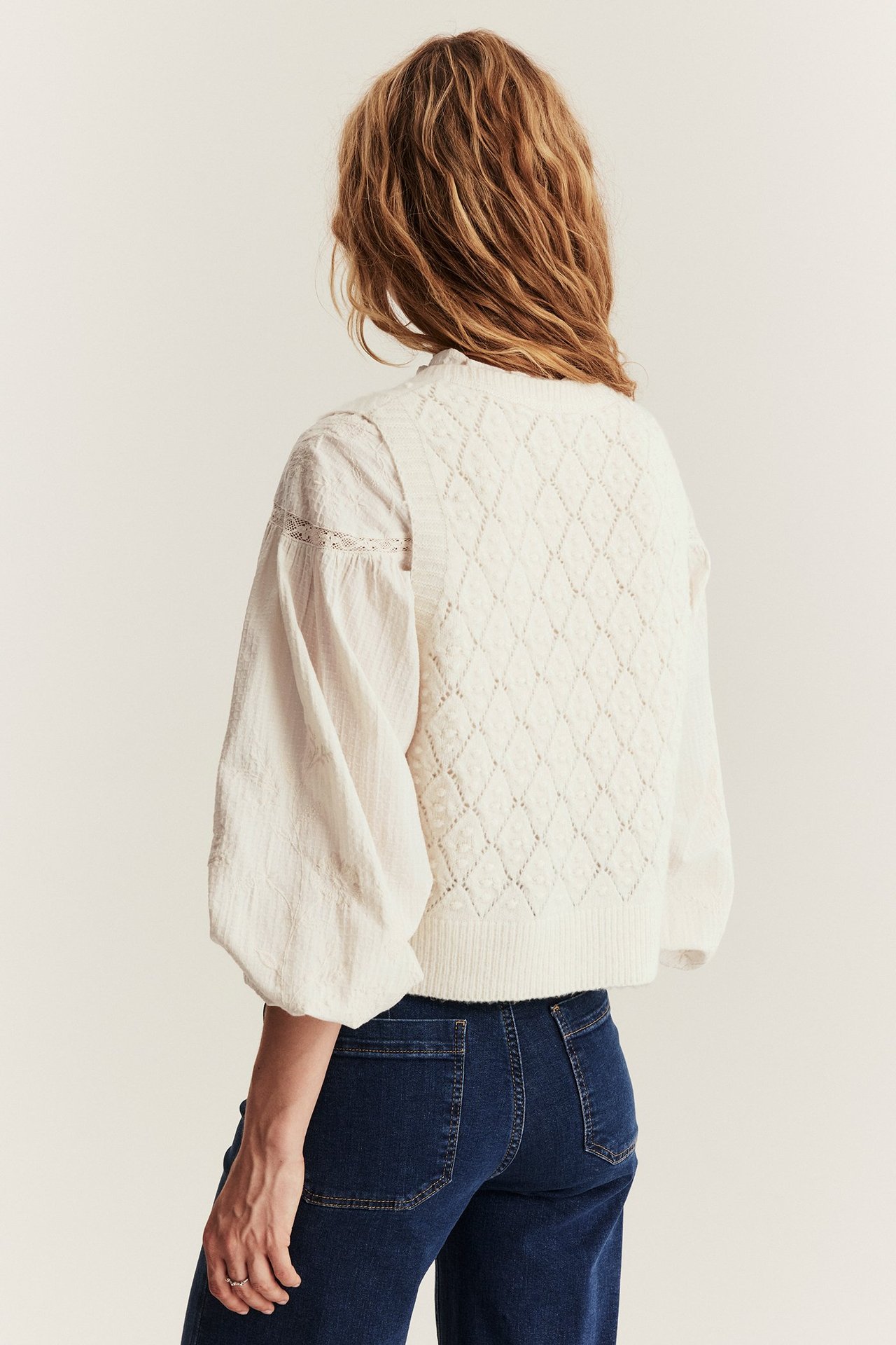 Back view of a woman wearing a light beige diamond knit wool blend vest over a white blouse and dark blue jeans.