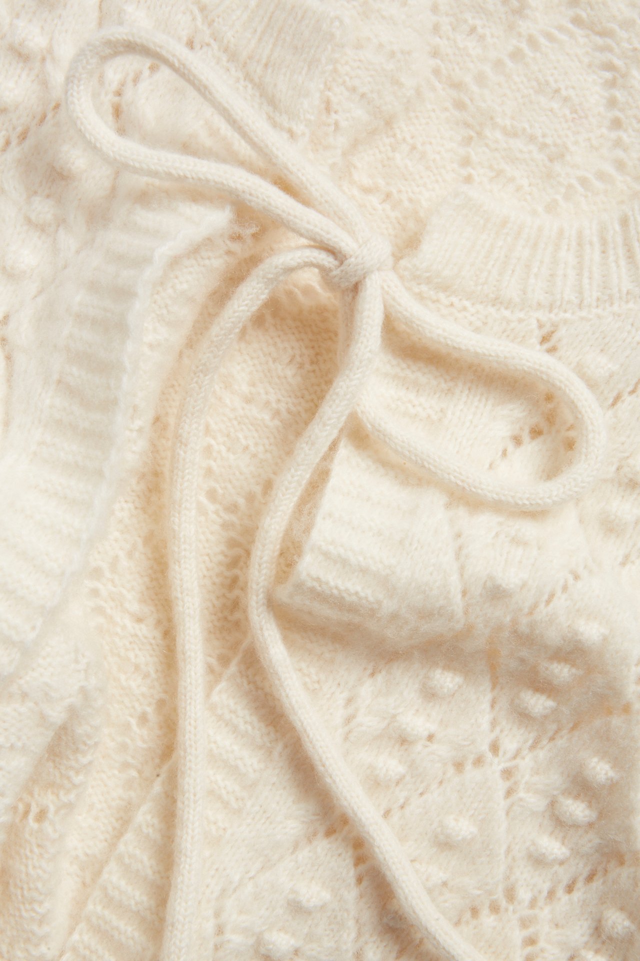 Close-up of a beige knitted wool blend vest with a tie-band and openwork pattern.