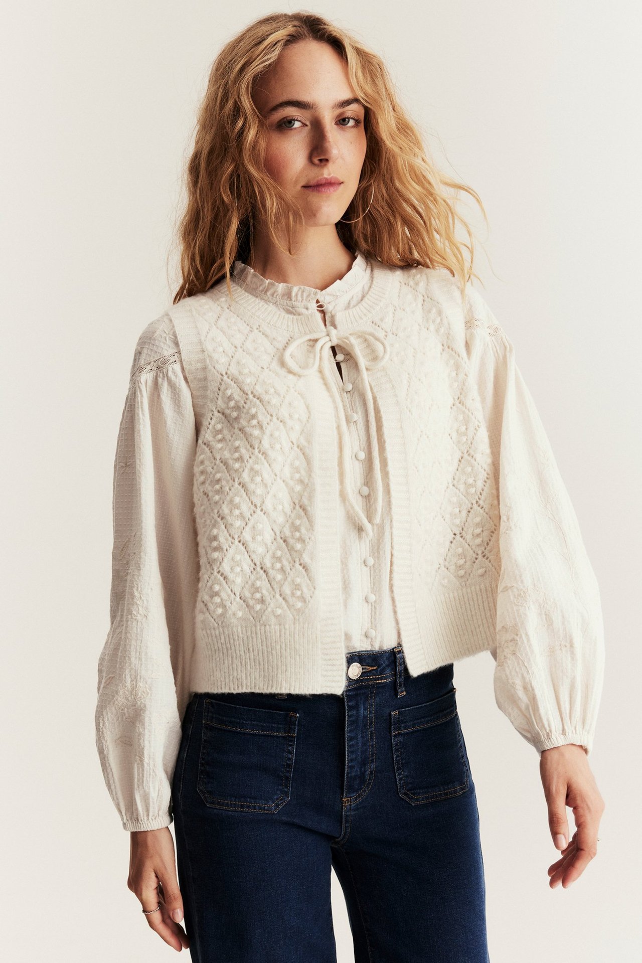 Front view: Woman in a beige melange wool blend vest with tie-front, white blouse, and dark blue jeans.